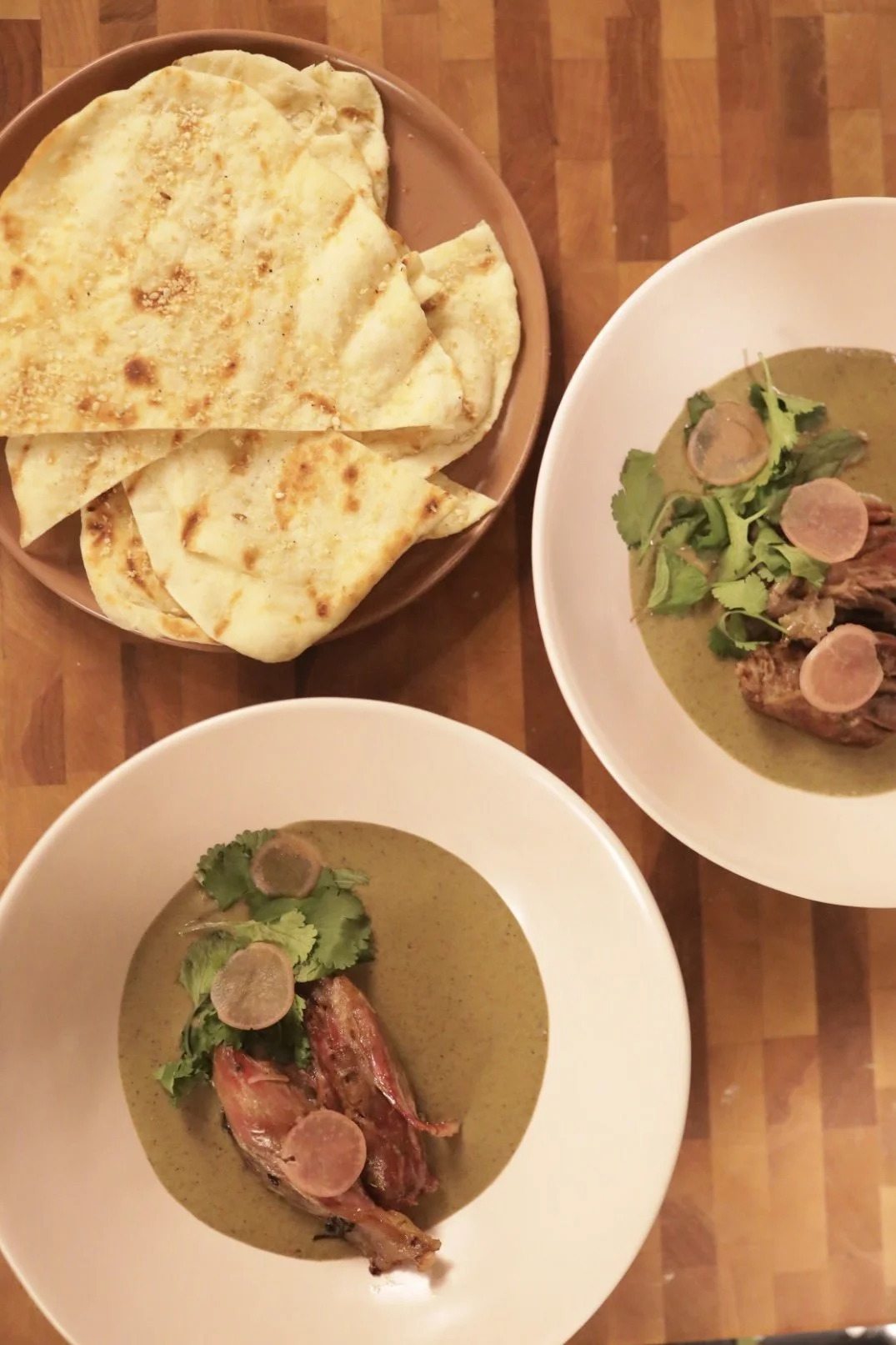 Two bowls of green curry with chicken, topped with sliced garlic, cilantro, and lime leaves, next to a small plate of flatbread or naan bread on a wooden surface.