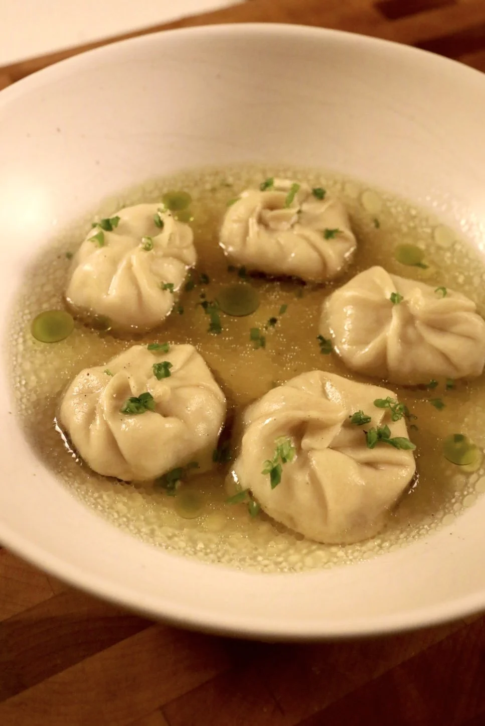 A white bowl with five steamed dumplings in broth garnished with chopped green herbs and small green peas.