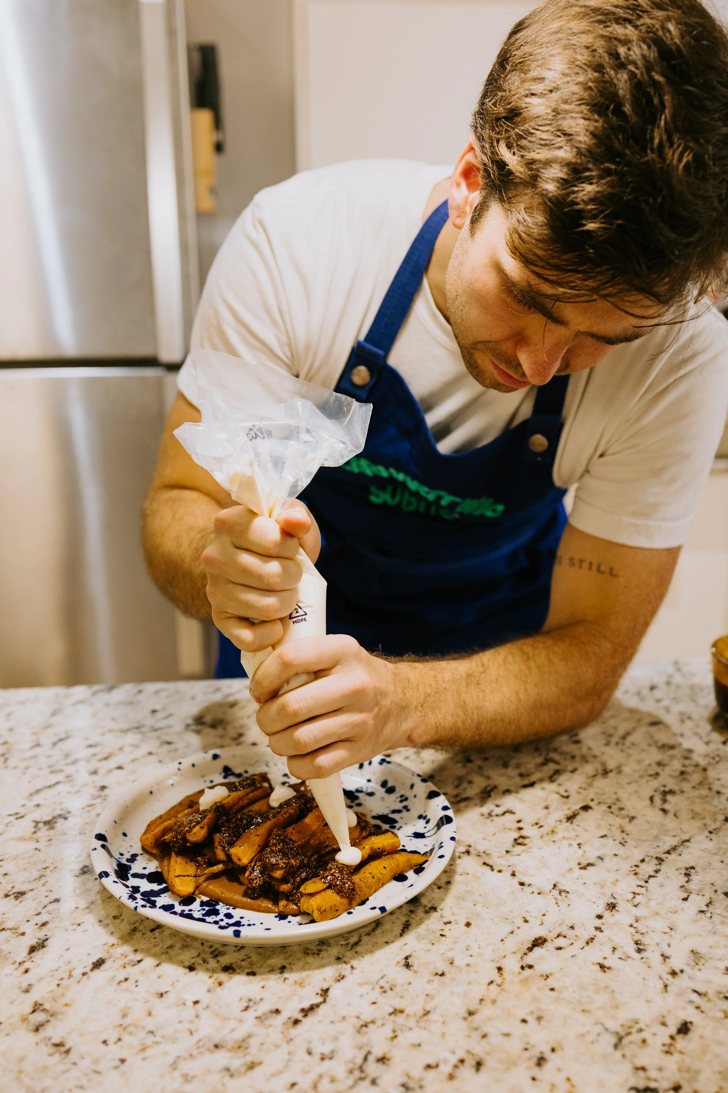 Man in a blue apron decorating a plate of food with piping bag in a kitchen.