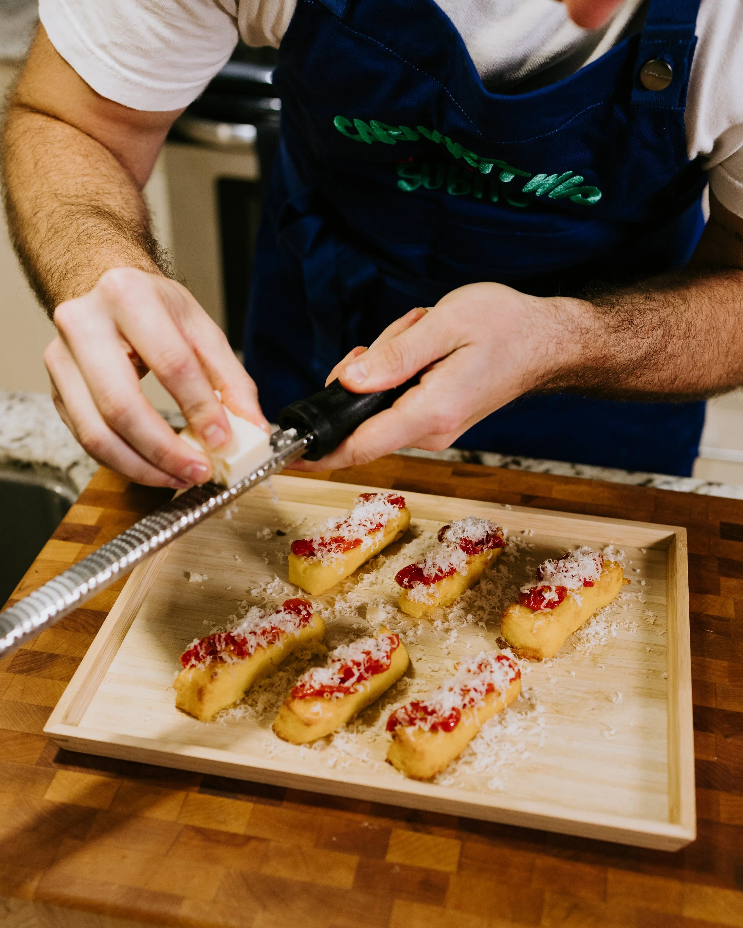 Person cutting cheese on a cheese grater, preparing finger foods topped with cheese and red sauce on a wooden tray.