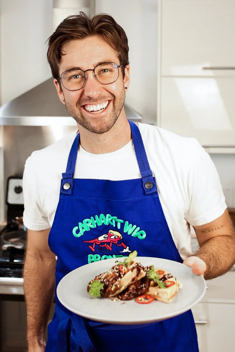 A smiling man wearing glasses, a white t-shirt, and a blue apron holding a plate of food in a kitchen.