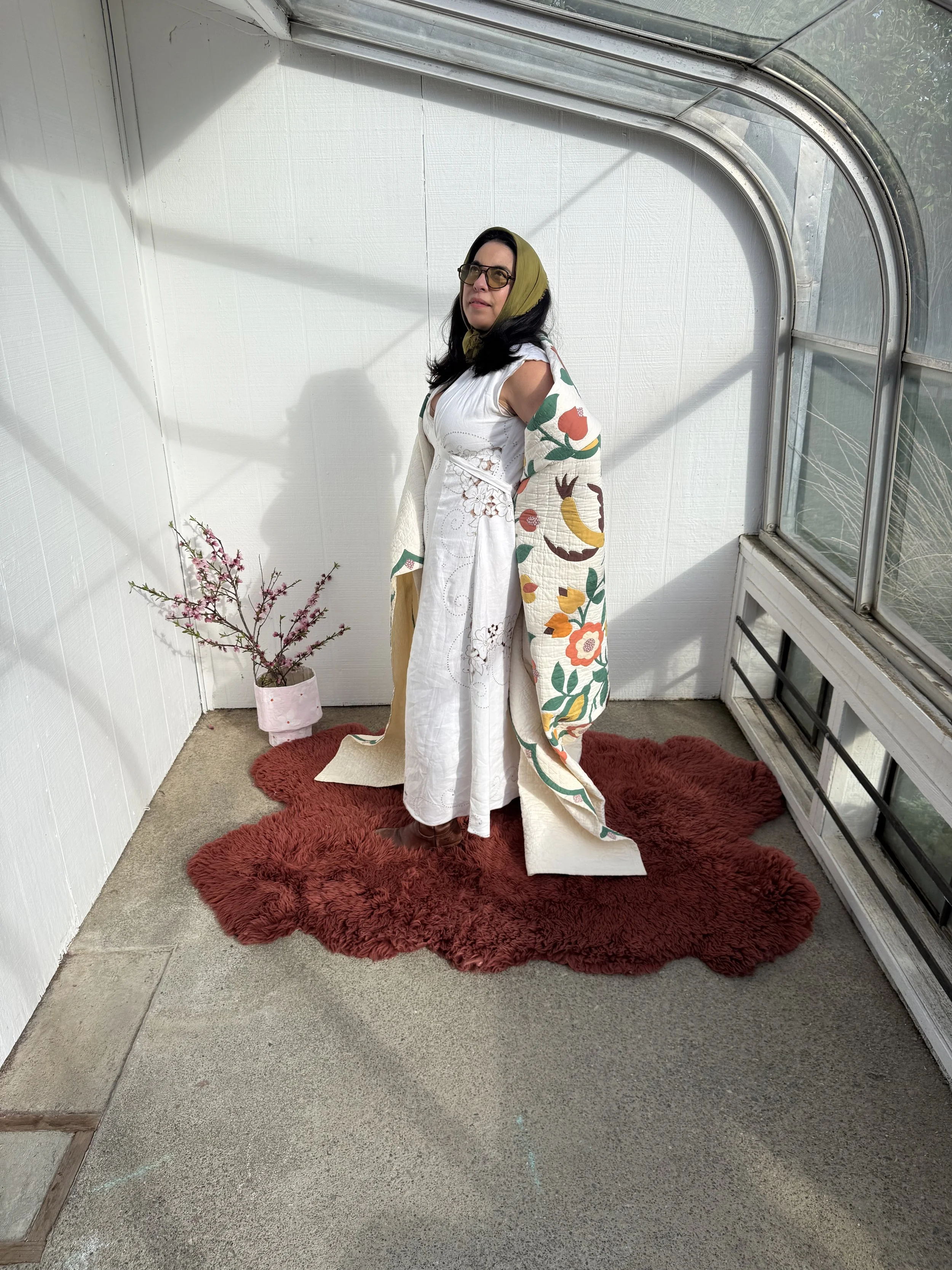 A woman wearing glasses, a headscarf, and a white dress, standing on a red furry rug in a bright greenhouse with white walls and large glass windows, next to a pink pot with a small flowering plant.