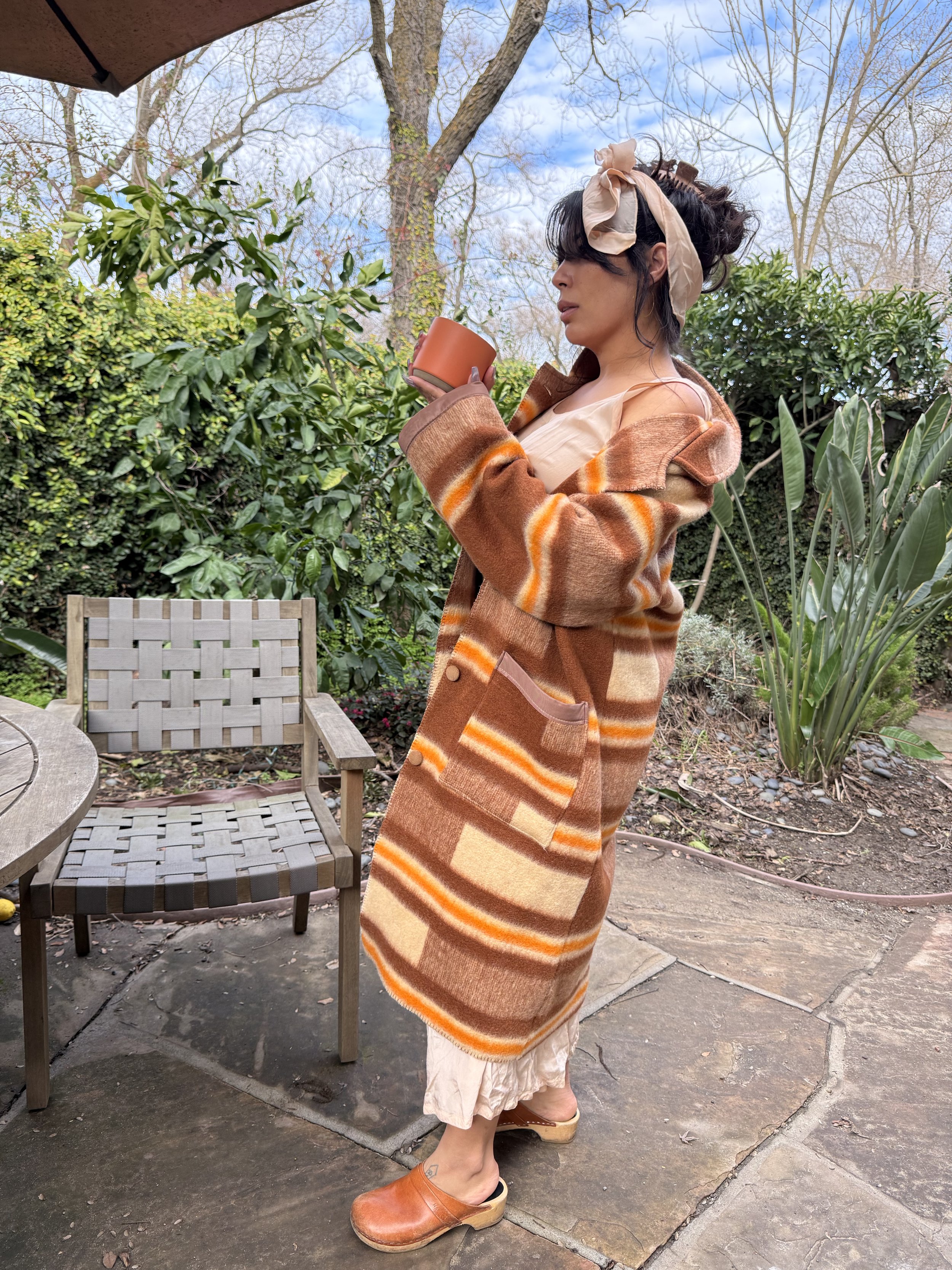Woman in a patterned coat and beige dress, holding a coffee mug, standing outdoors in a garden with trees and plants.