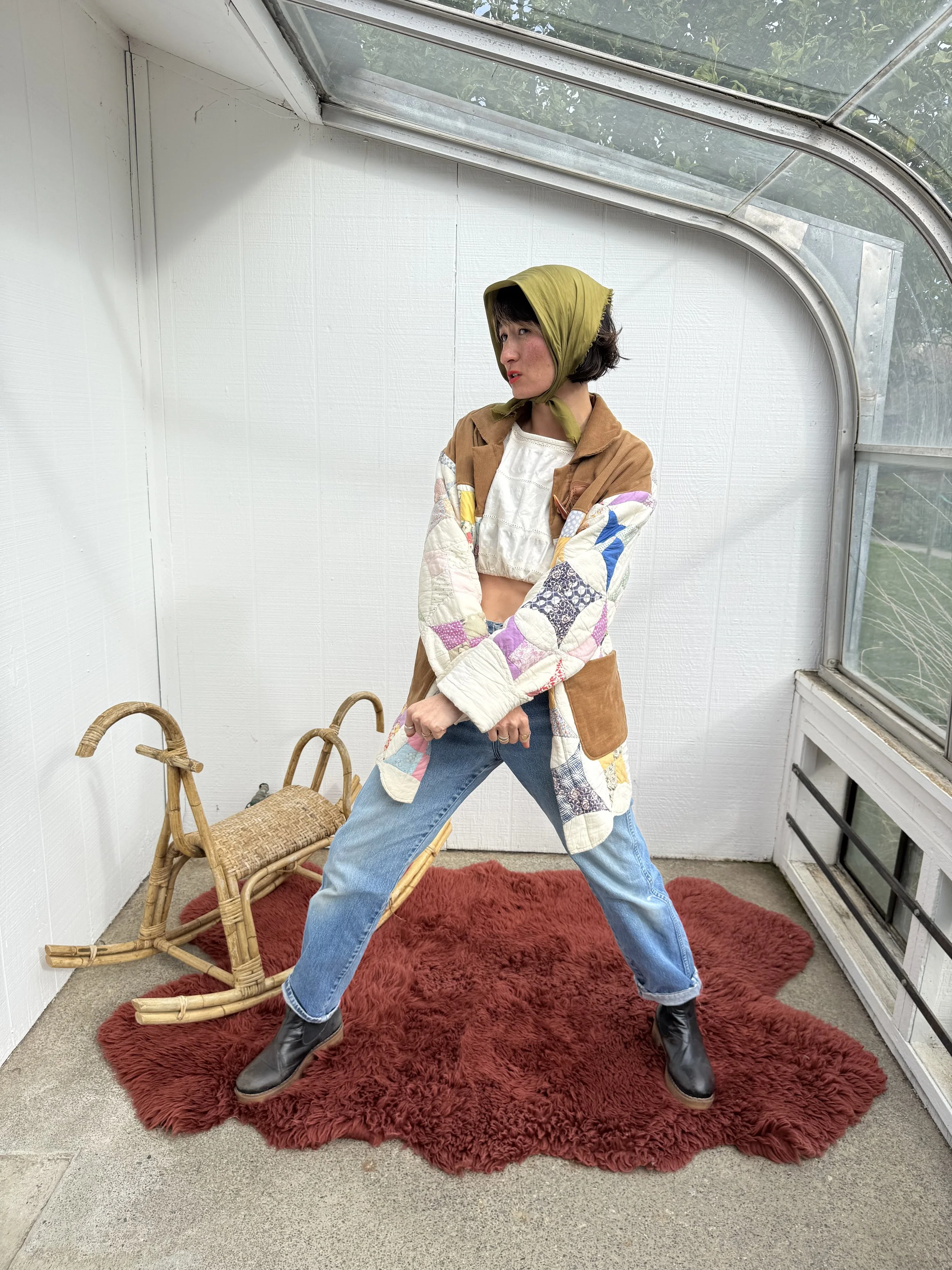 A woman wearing a yellow headscarf, a patchwork coat, a crop top, and jeans stands in a greenhouse corner on a red shaggy rug, with a vintage wooden sled beside her.