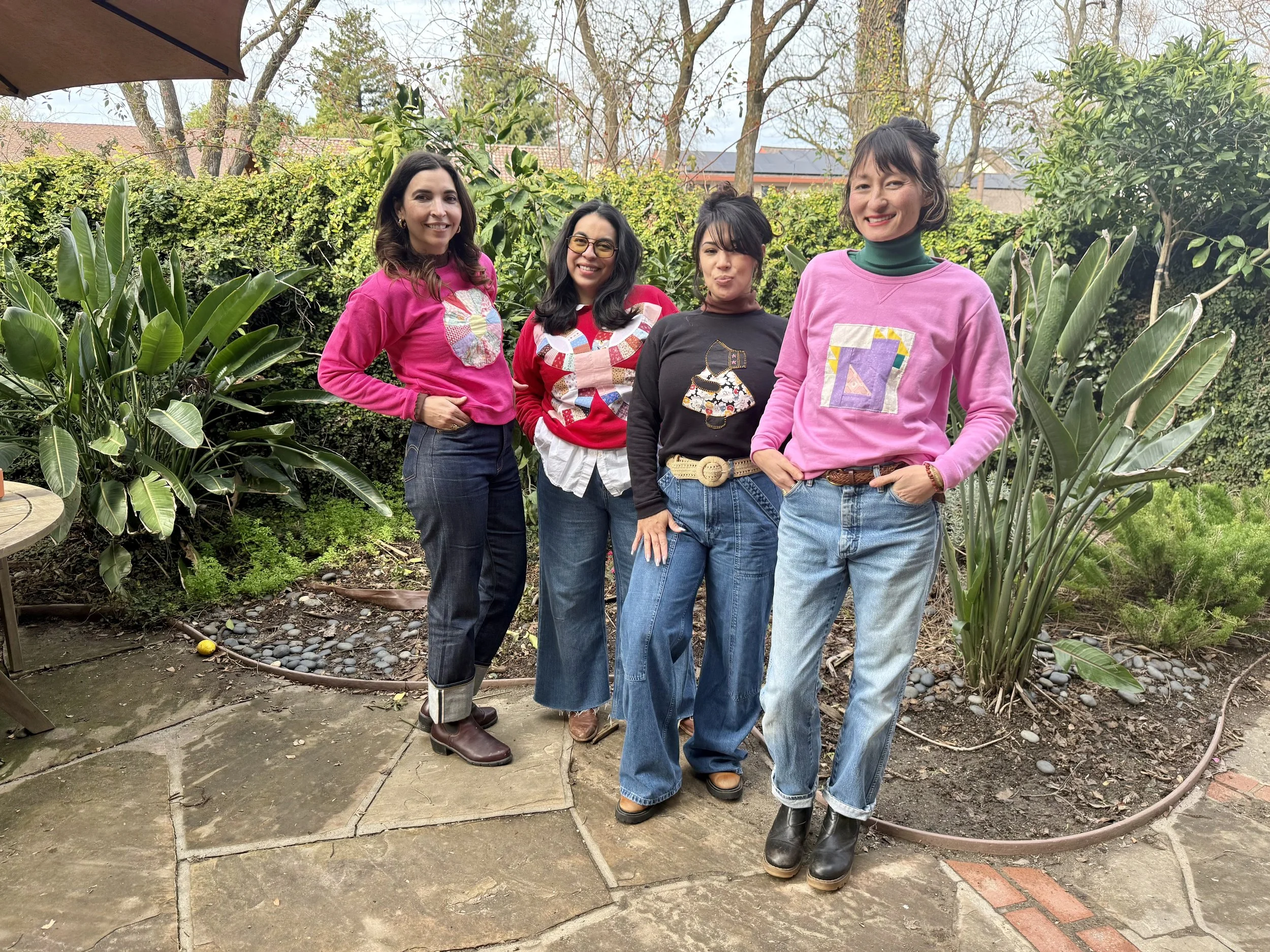 Four women standing outdoors on a stone patio, smiling at the camera, with greenery and trees in the background. They are wearing colorful sweaters with artistic designs.