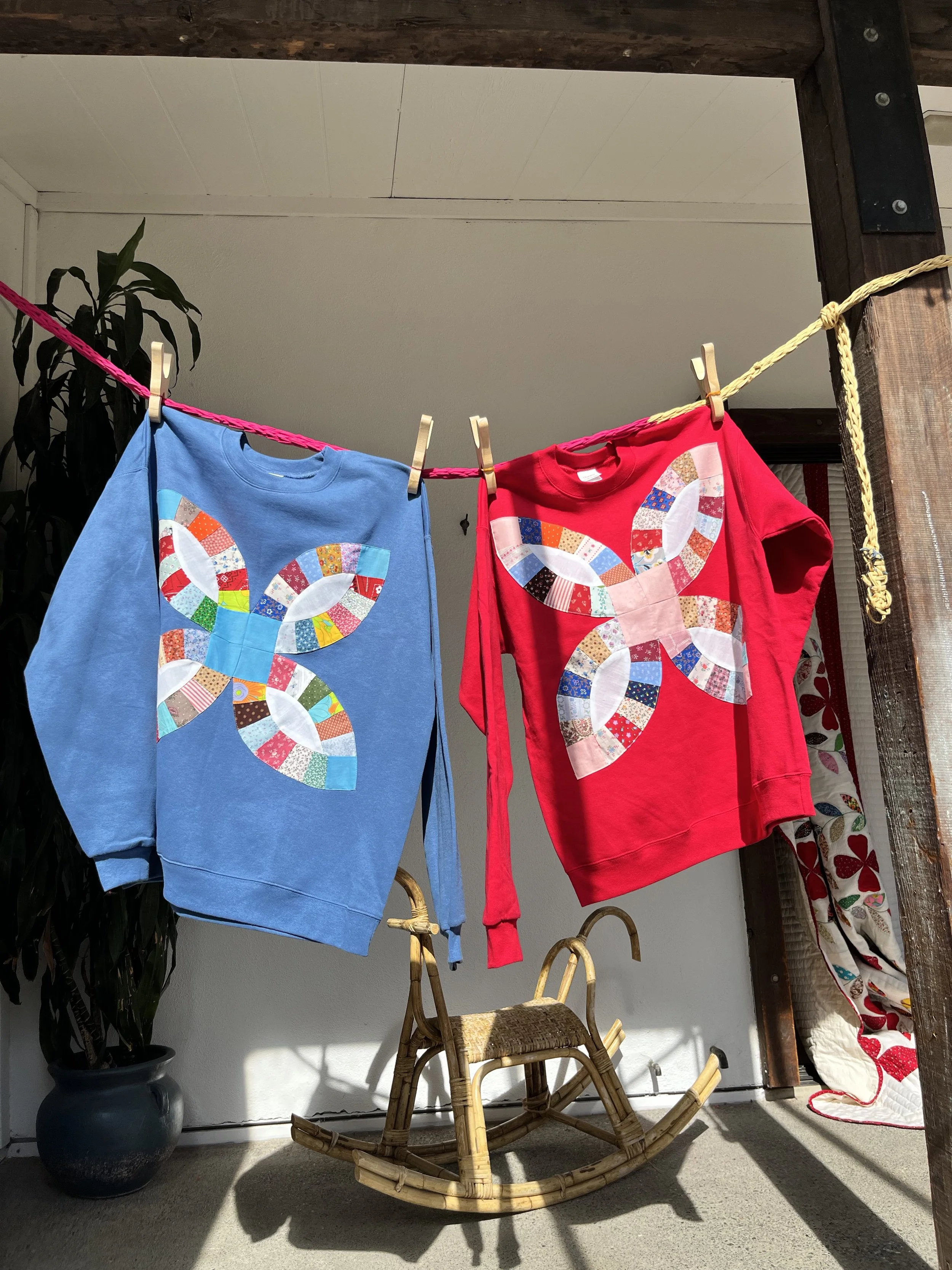Two sweatshirts hanging on a pink and yellow rope, each with a colorful butterfly design on the front, hanging from wooden clothespins. There is a rocking chair and a potted plant in the background.