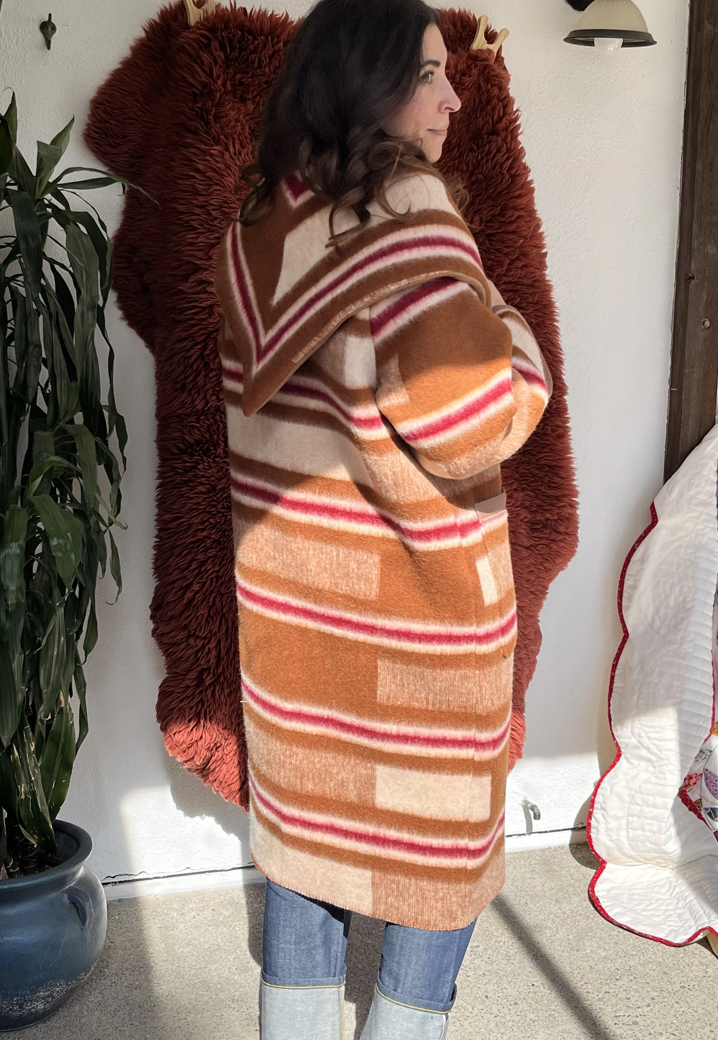 A woman with dark wavy hair standing inside a room, wearing a multicolored striped sweater with shades of orange, red, cream, and brown. She is standing in front of a large, fluffy, red blanket or wall hanging, with her arms crossed, looking to the r