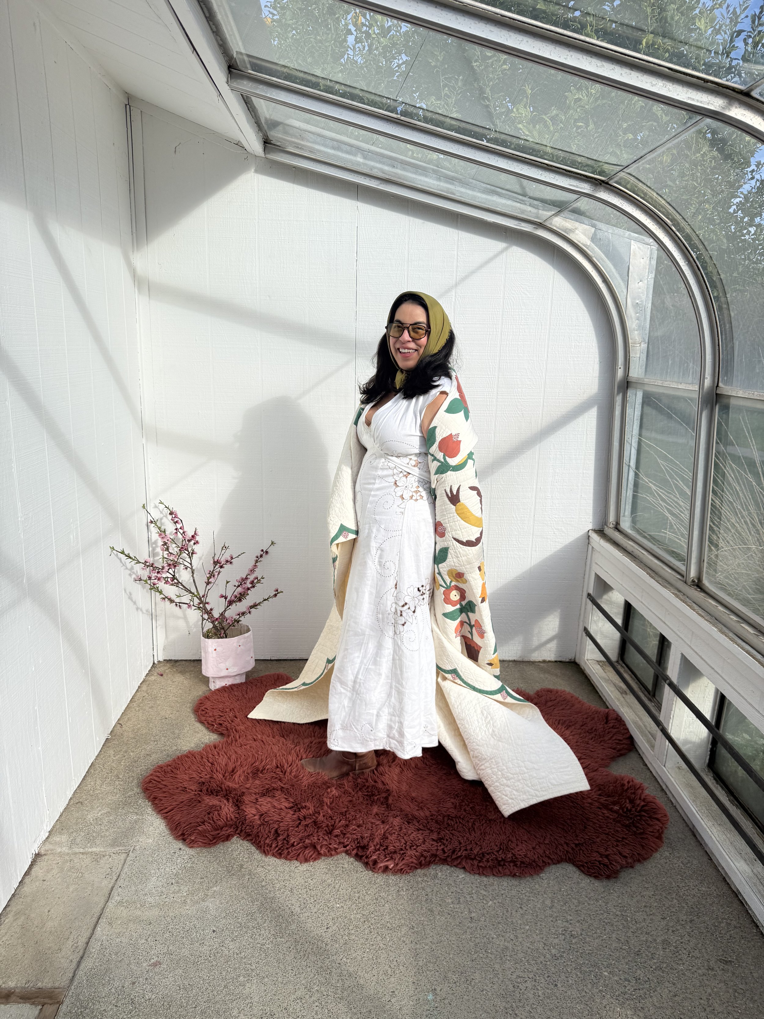 A woman with dark hair, glasses, and a green headscarf, standing on a red rug inside a white room with a large glass window. She is wearing a white dress with colorful floral-patterned sleeves and smiling at the camera.
