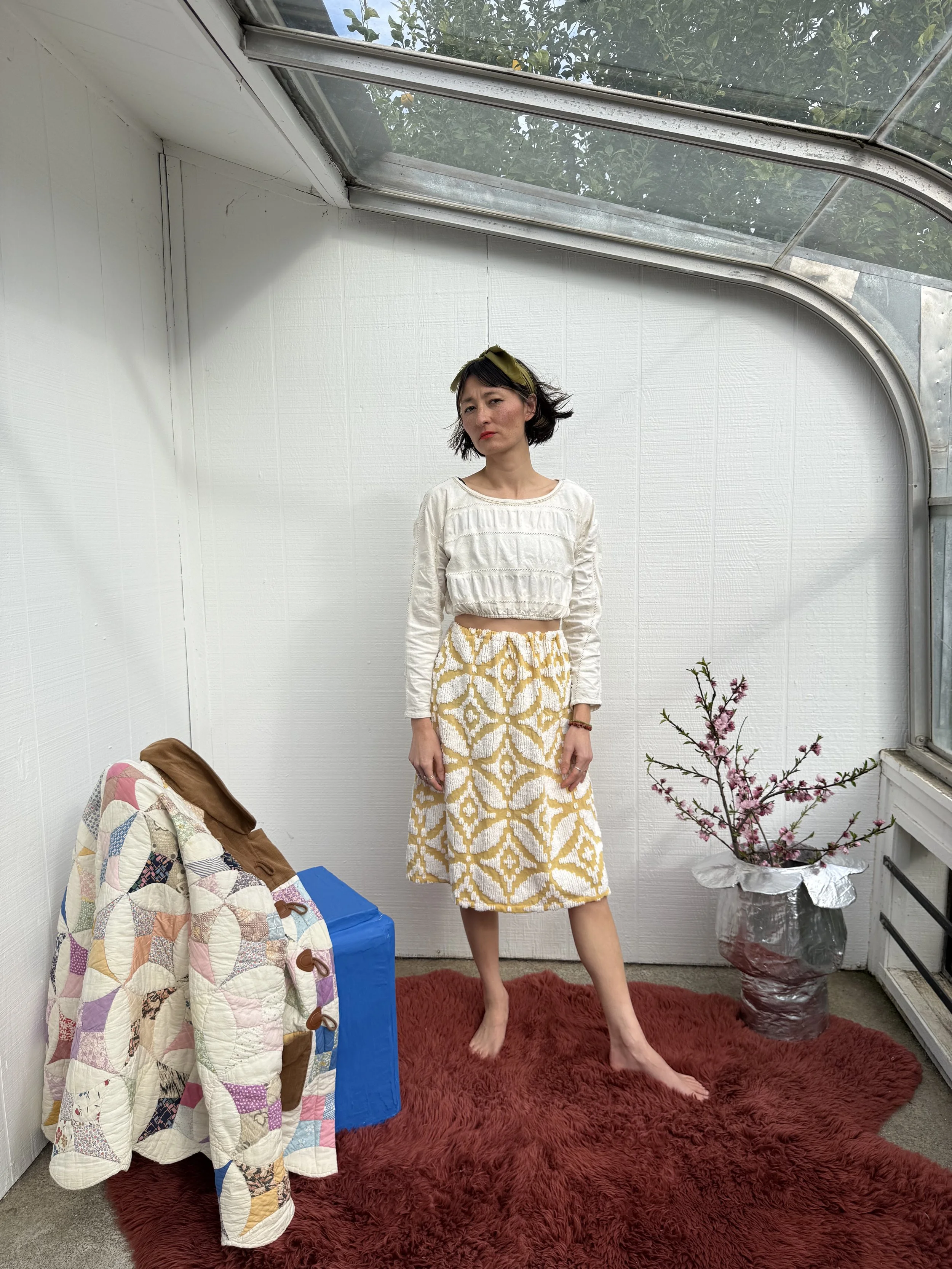 A woman with short black hair and a green headband stands barefoot on a red furry rug in a white-paneled indoor space with a skylight ceiling. She wears a cream-colored long-sleeve crop top and a yellow and white patterned knee-length skirt. To her l