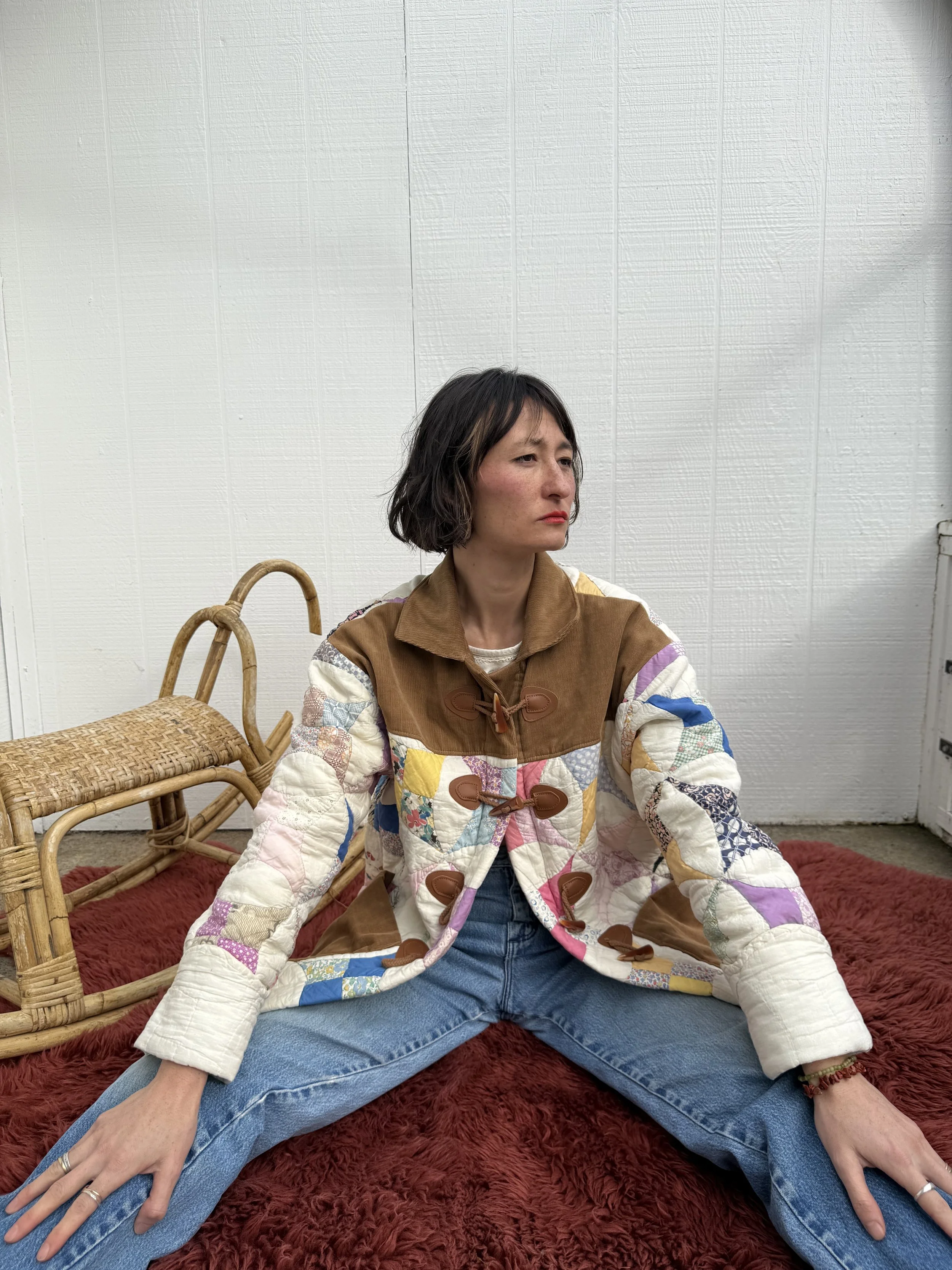 Woman sitting on a red furry rug, wearing a colorful patchwork quilted jacket and blue jeans, with a wicker chair beside her and a plain white wall in the background.