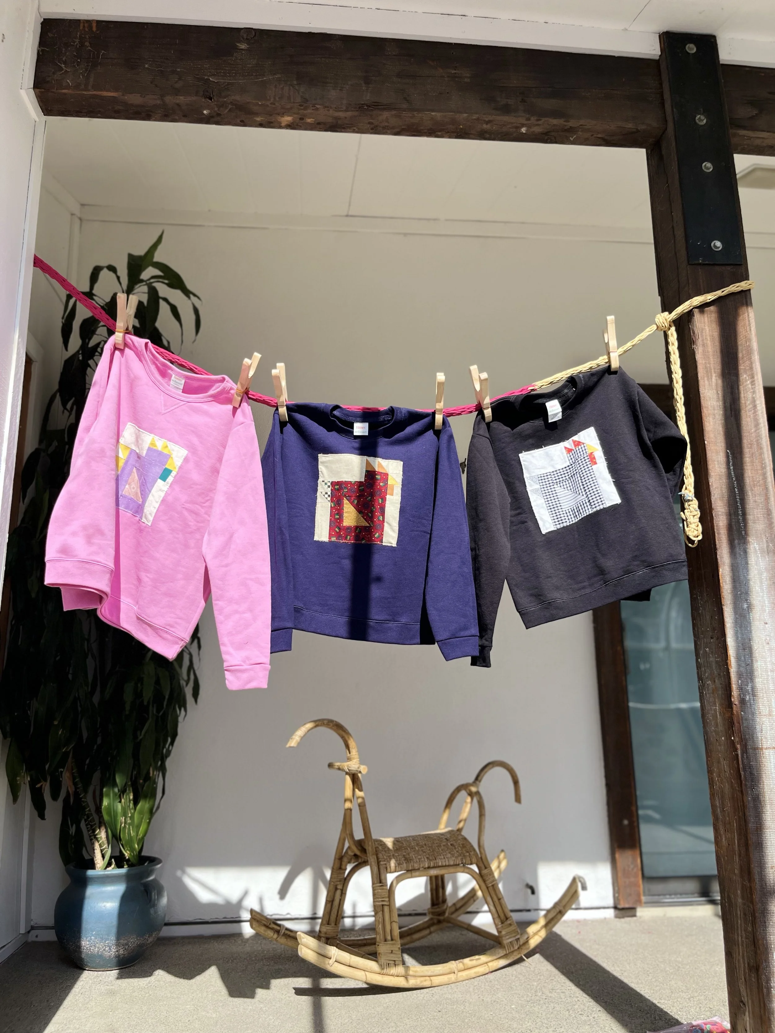 Three small sweatshirts hanging on a clothesline outdoors. The shirts are pink, purple, and black, each with colorful patch designs. Behind the shirts, there is a potted plant and a bamboo rocking horse on the ground.