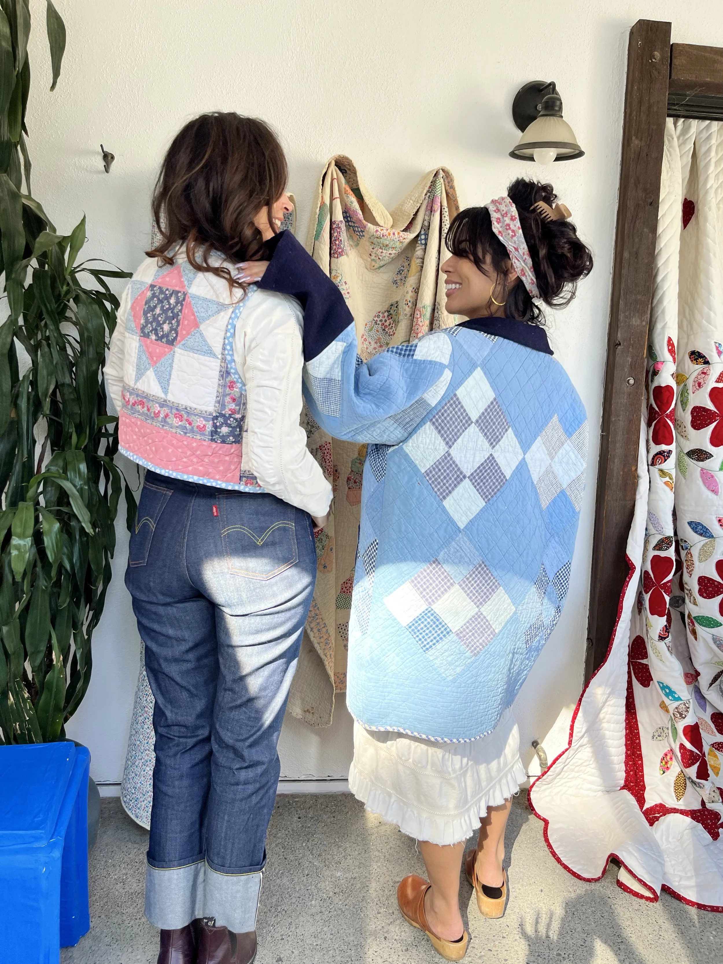 Two women are standing and talking in front of a wall of quilts. They are wearing colorful, patchwork-style jackets and are smiling at each other.