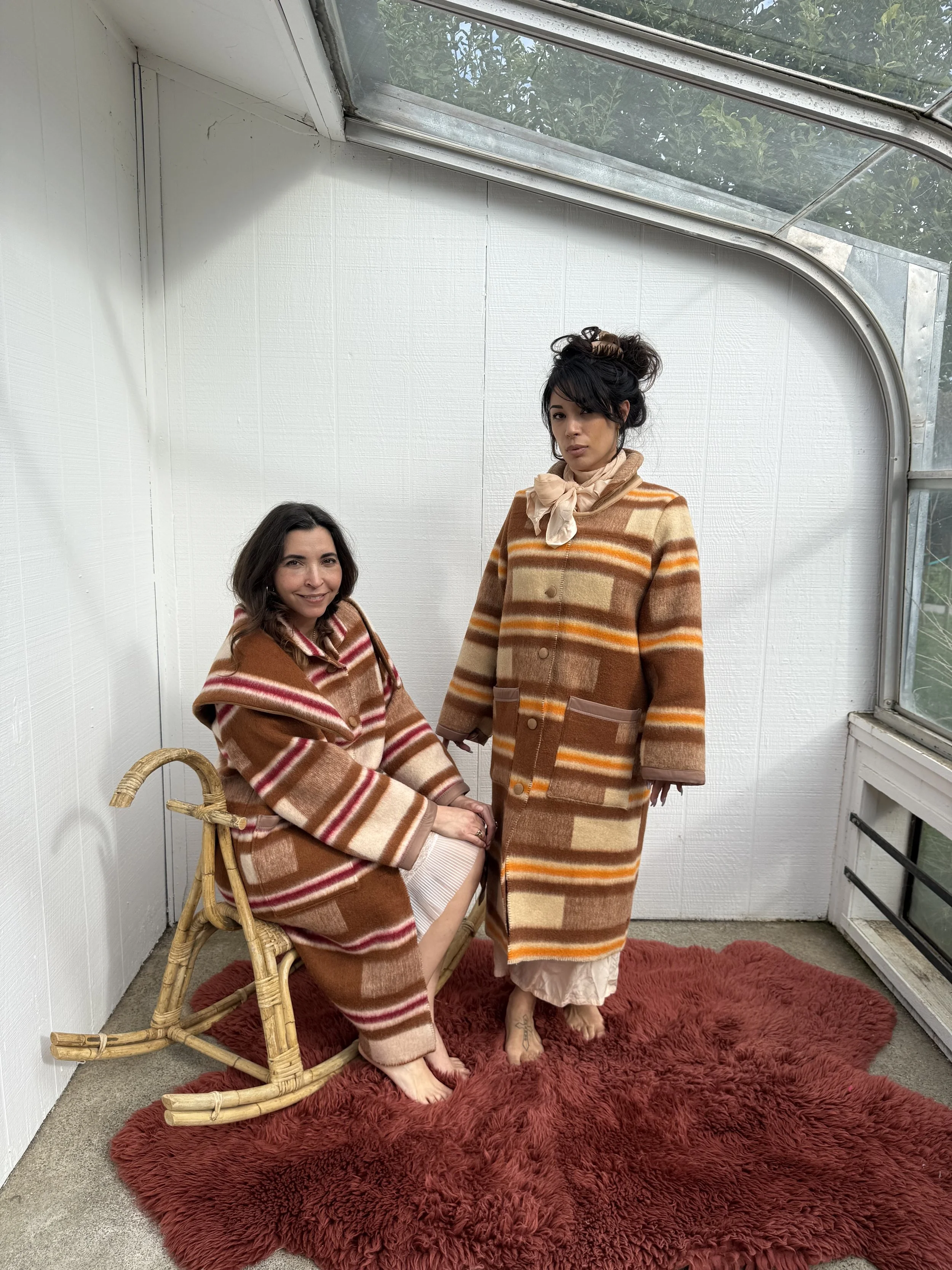 Two women wearing brown, orange, and beige striped coats in a sunroom with a glass ceiling and window. One woman is seated on a wicker chair, smiling, and the other is standing, looking serious. They are barefoot on a red shaggy rug.