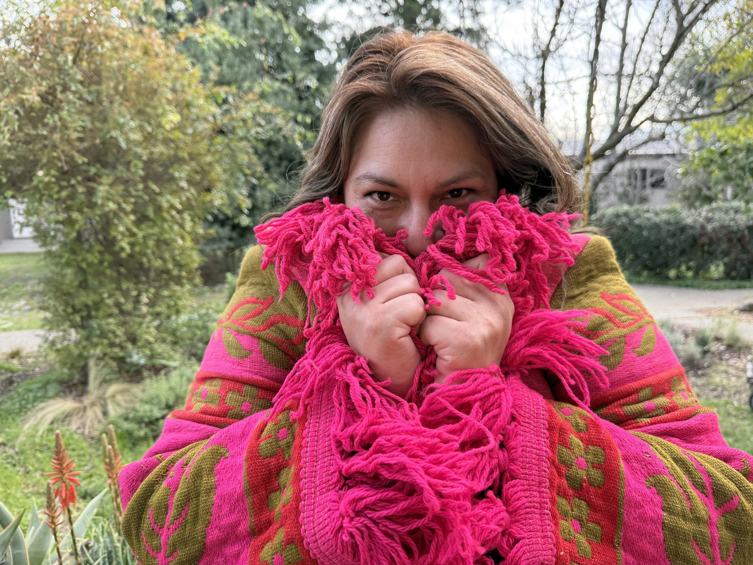 Woman with brown hair, holding a bright pink, knitted scarf up to her face, peeking over it with her eyes smiling, outdoors in a garden with trees and plants.
