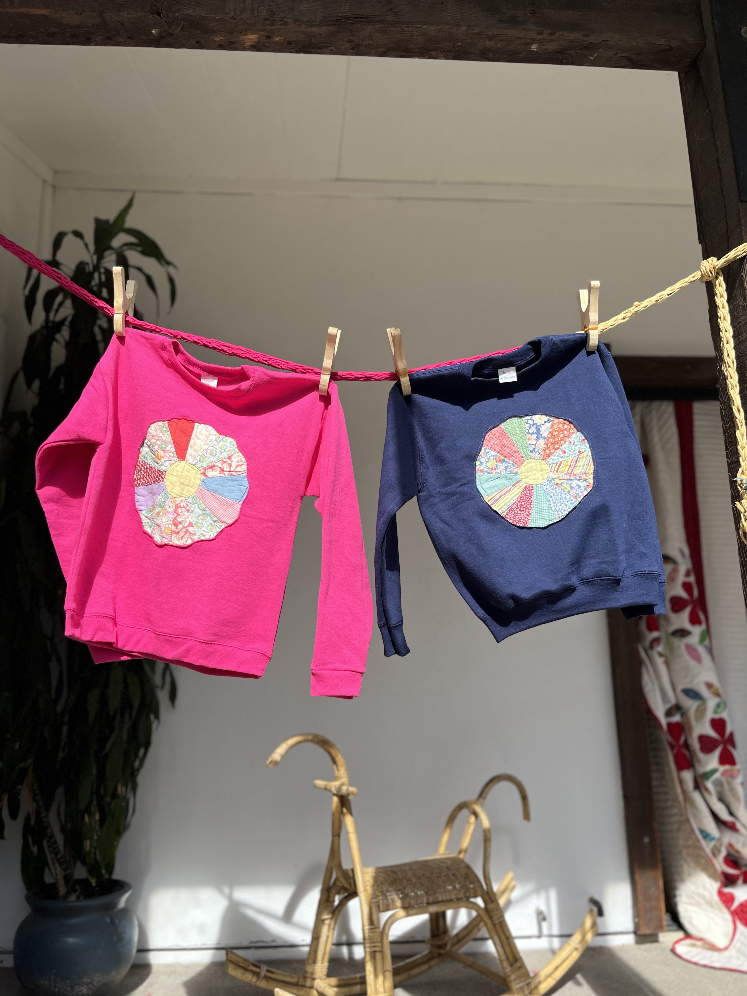 Two children's sweatshirts, one pink and one blue, hanging on a clothesline with clothespins. Each sweatshirt has a multicolored patch in the shape of a flower on the front.