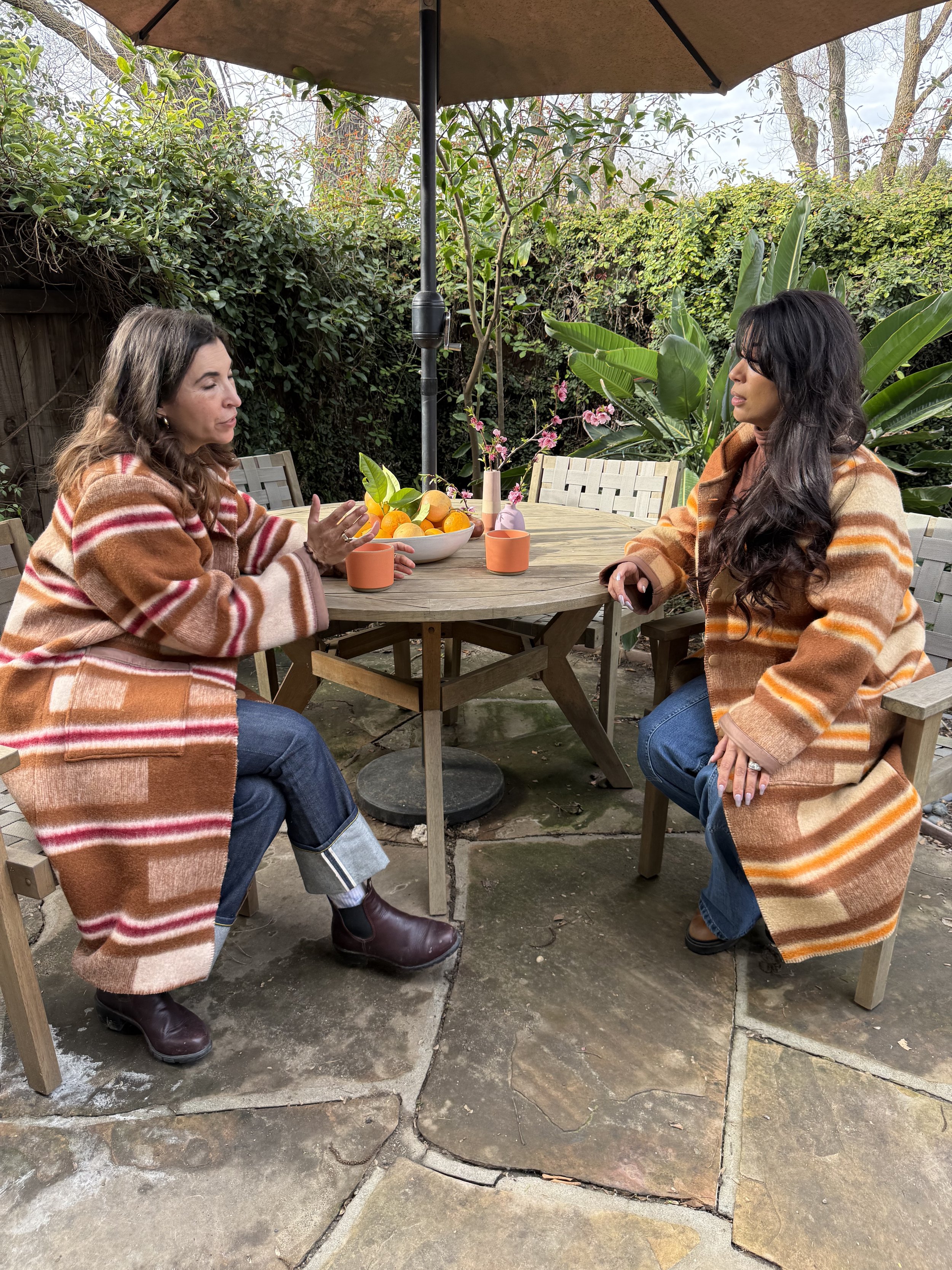Two women sitting at a wooden patio table engaged in conversation outdoors, with a large umbrella overhead and lush greenery in the background, decorated with a bowl of oranges, pink flowers, and candles.