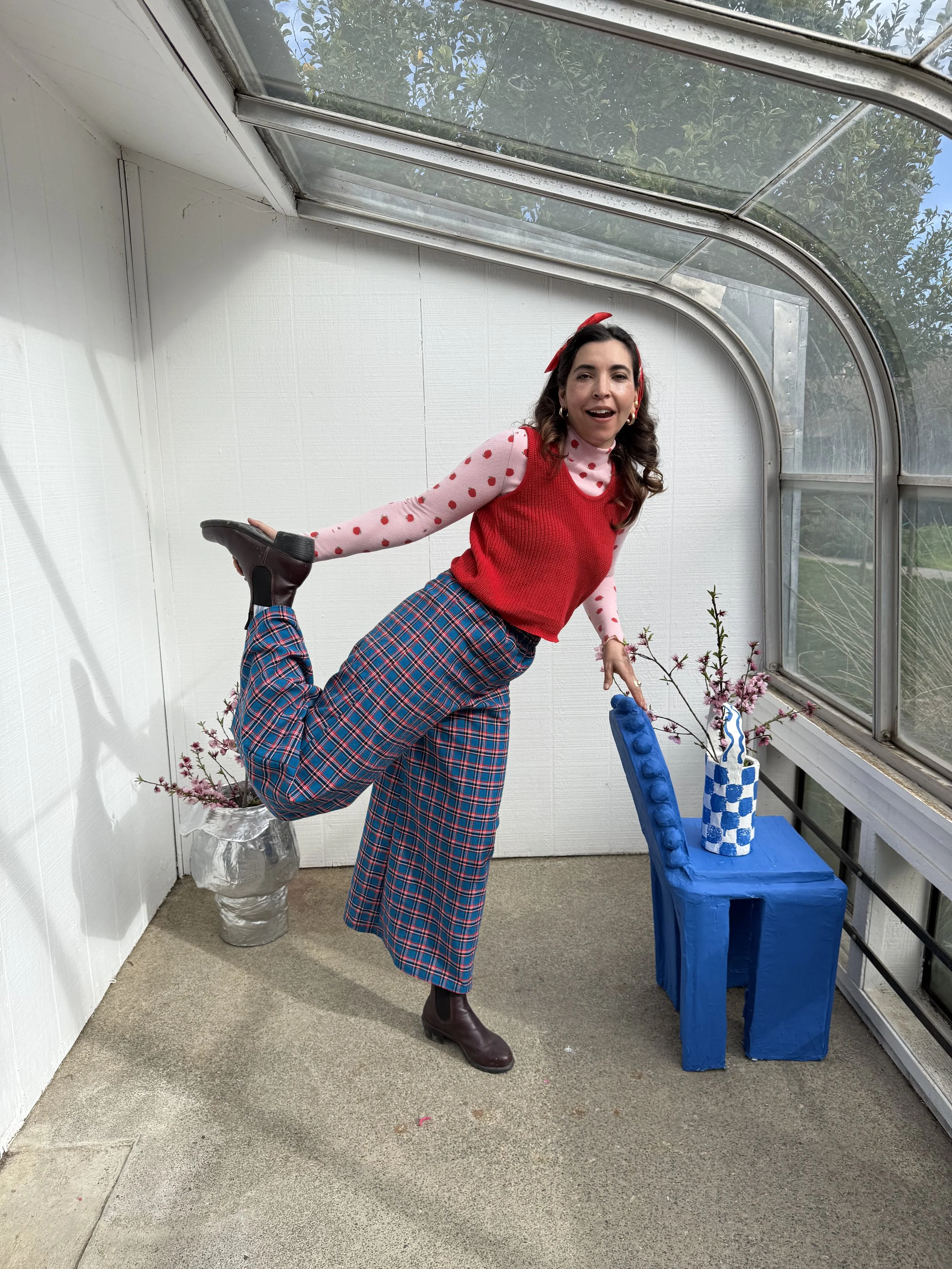 A woman wearing a red vest, pink polka-dot shirt, plaid wide-leg pants, and brown boots is standing in a glass-enclosed space, holding her left ankle behind her with her left hand, while her right hand is resting on a blue table with a checkered blue