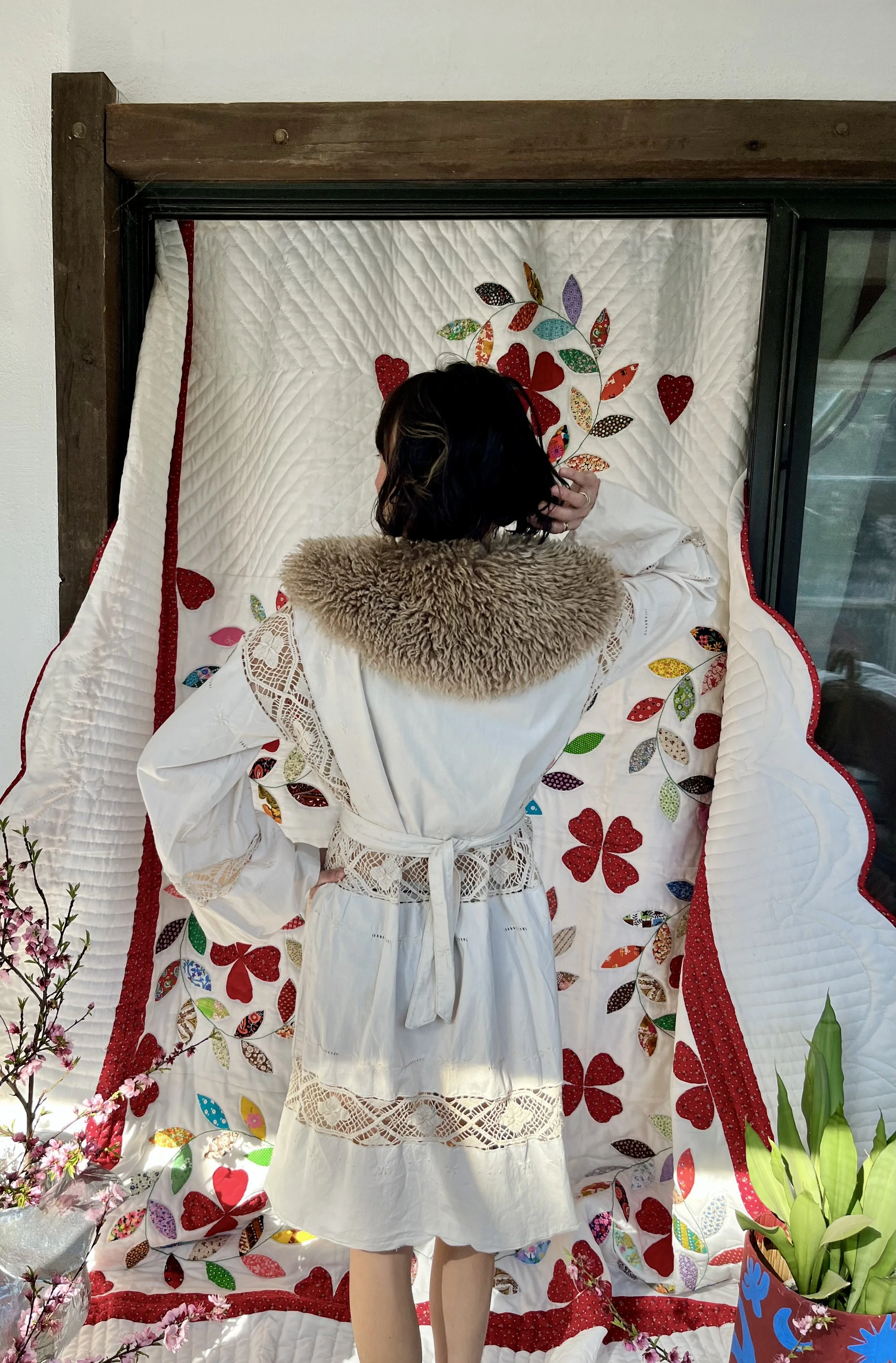 A woman with black hair and a beige fur collar, wearing a white dress with lace details, standing in front of a quilted wall hanging decorated with colorful fabric hearts and leaves.