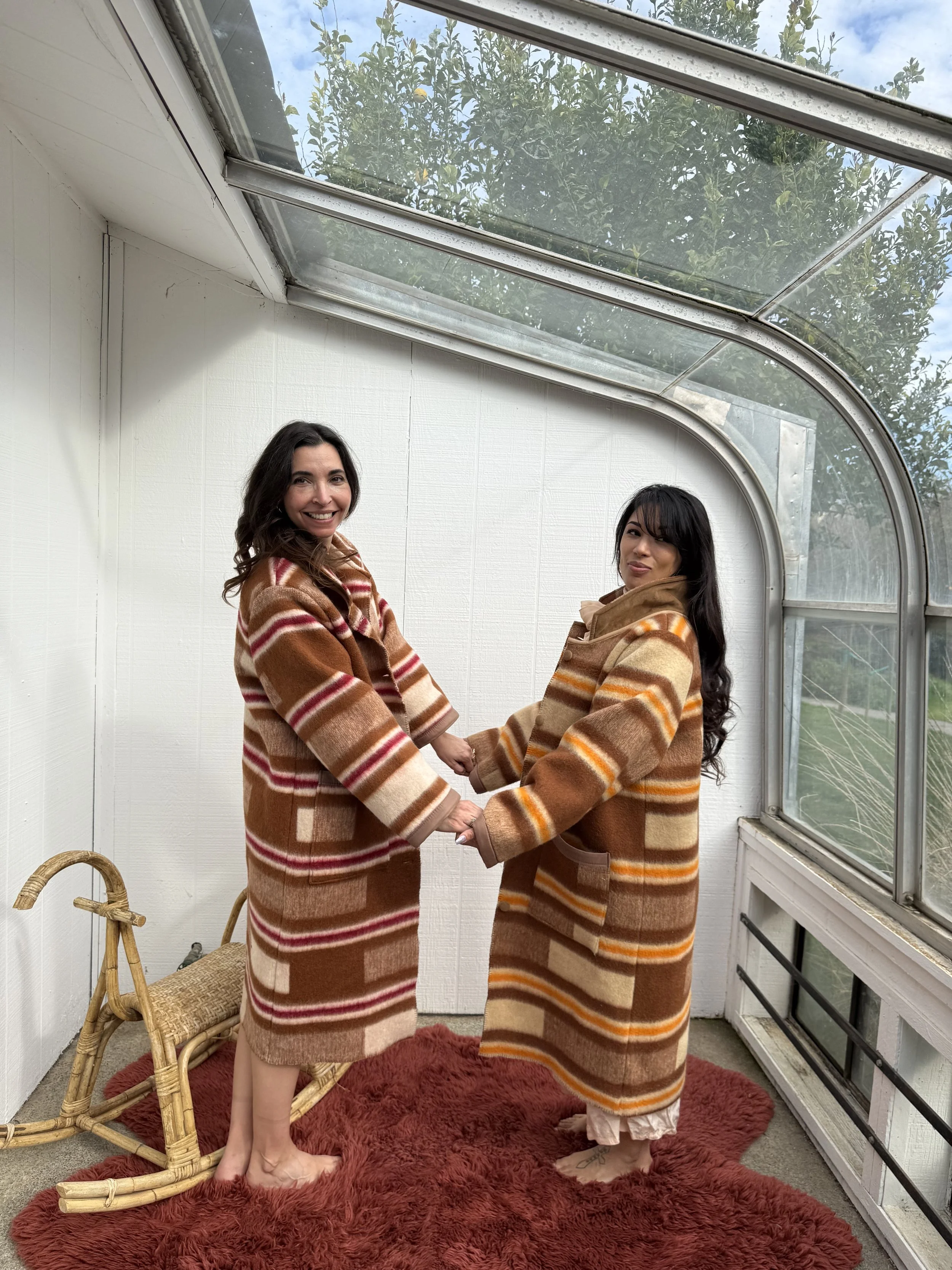 Two women holding hands and wearing matching brown plaid coats inside a sunroom with large windows, a red rug, and a wicker bench.