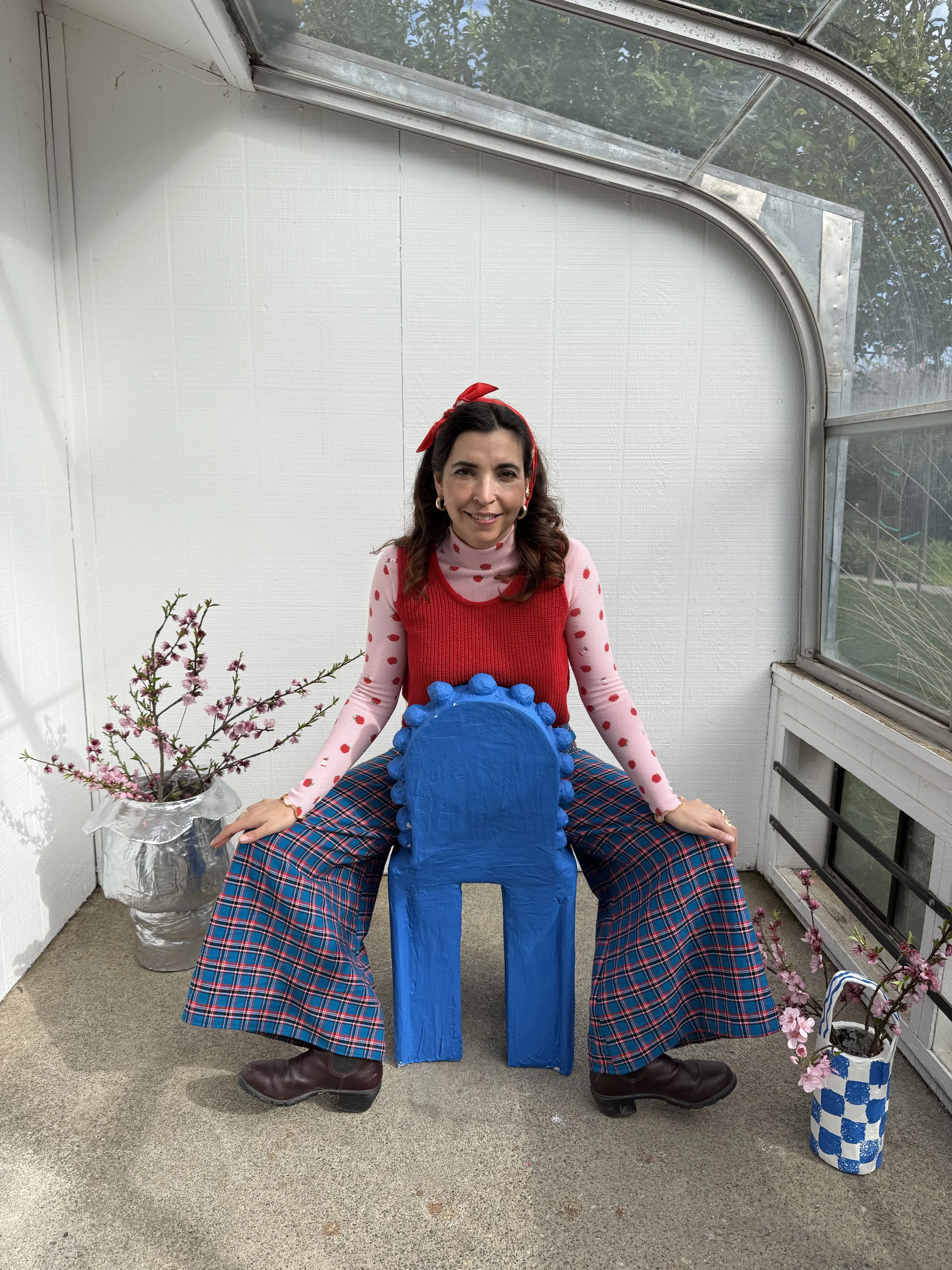 Woman dressed in red and pink with plaid pants, sitting on a blue, painted wooden chair, inside a sunroom with potted pink flowers on either side.