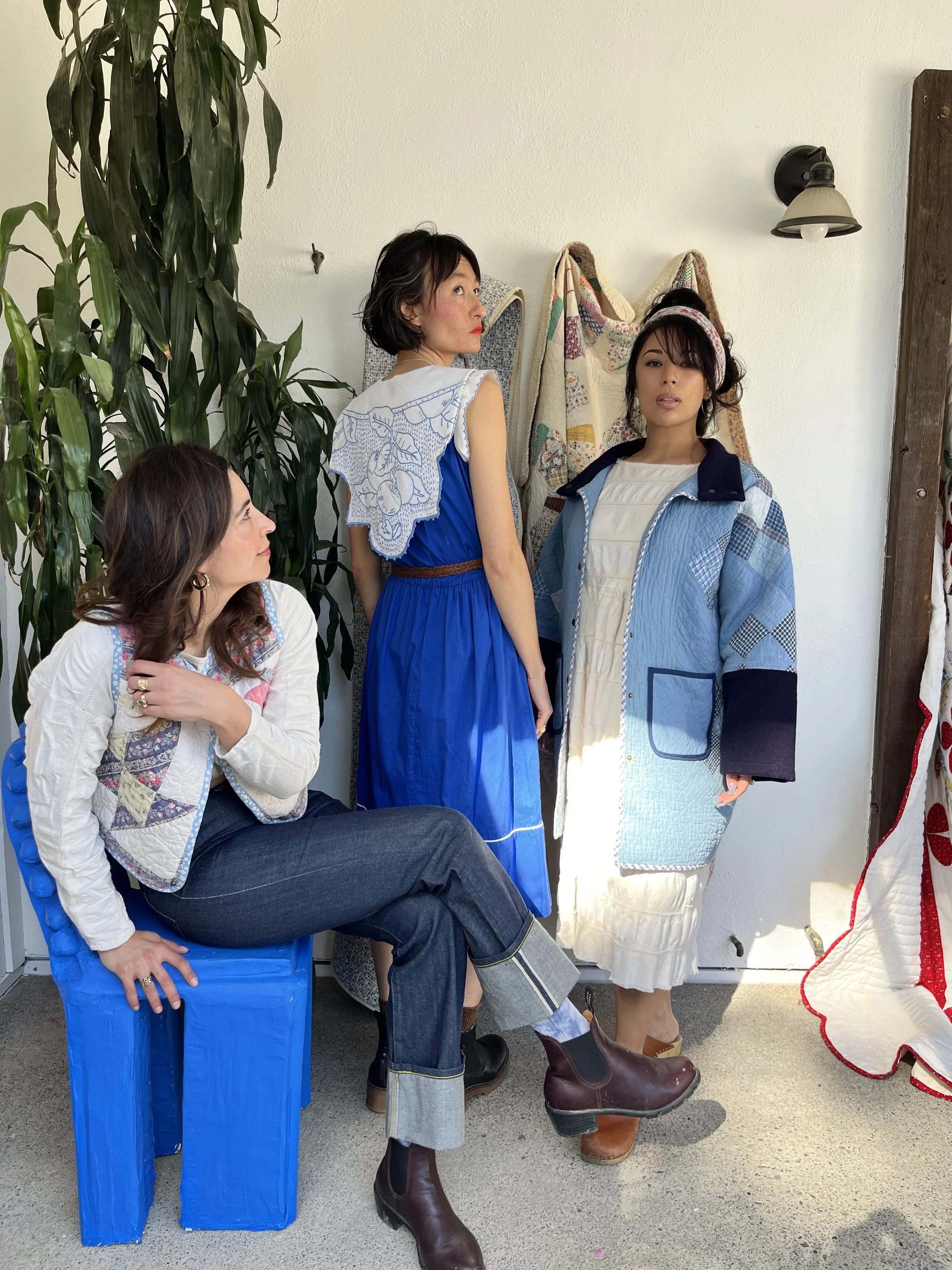 Three women with dark hair posing inside a room with a large green plant and quilted textiles hanging on the wall. One woman is sitting on a blue plastic chair, another standing with her back partially turned, and the third standing in front, wearing