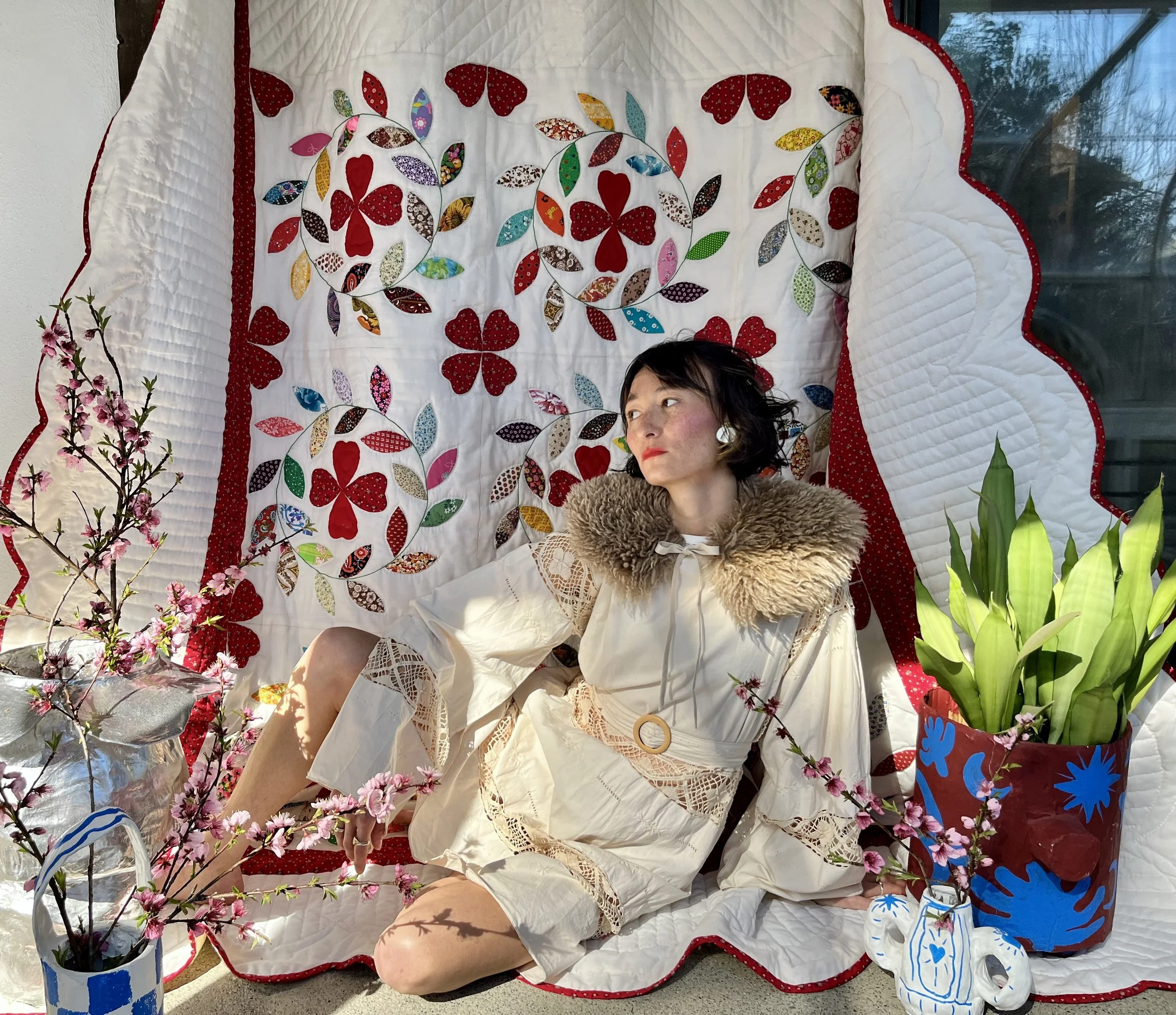 A woman with dark hair and fair skin sitting on a quilted backdrop with red and white patterns, surrounded by colorful pottery and pink flowering branches.