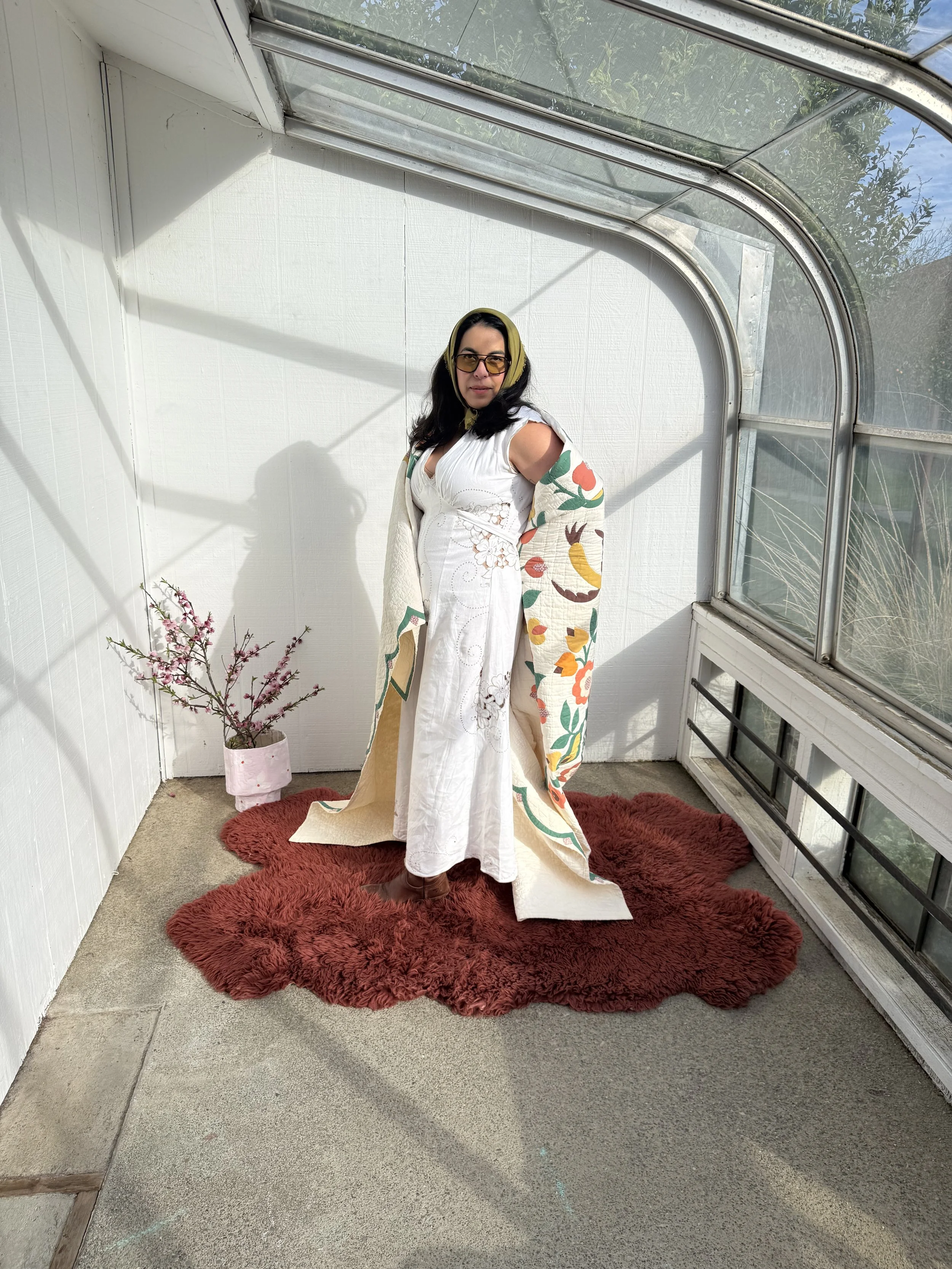 A woman in a white dress with embroidered floral patterns and a colorful embroidered shawl, standing on a red shaggy rug inside a sunlit greenhouse with large windows and a small pink flower pot with branches inside.
