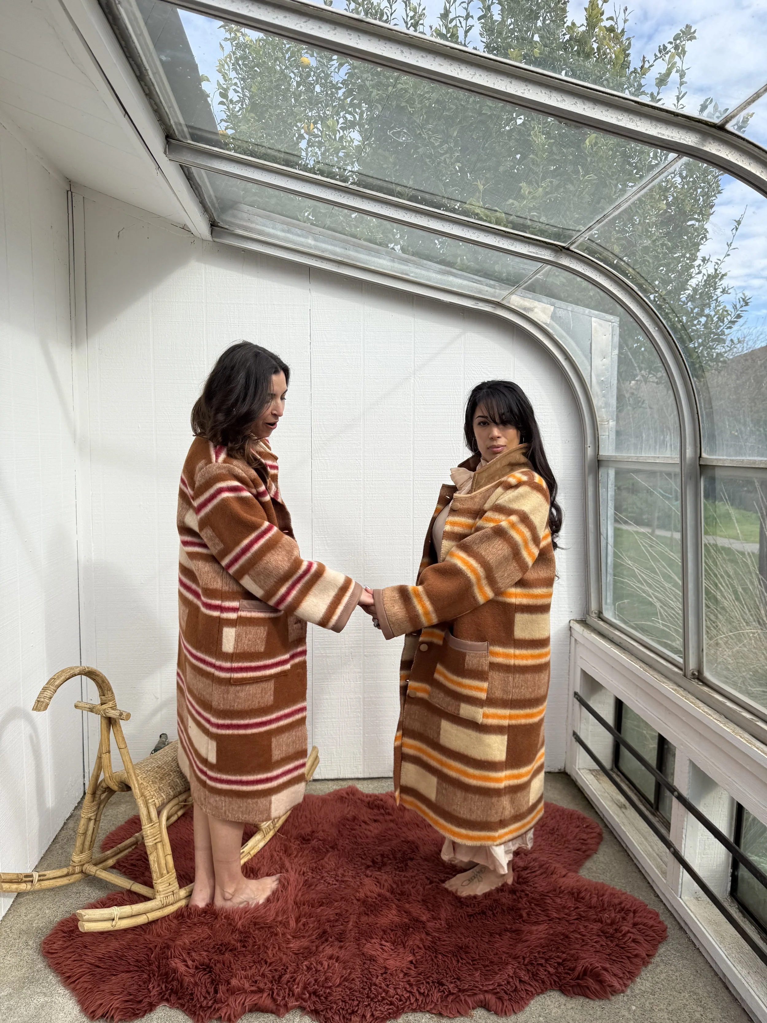 Two women wearing matching brown, orange, and cream striped coats facing each other and holding hands inside a glass greenhouse with a red shaggy rug and a wooden chair.