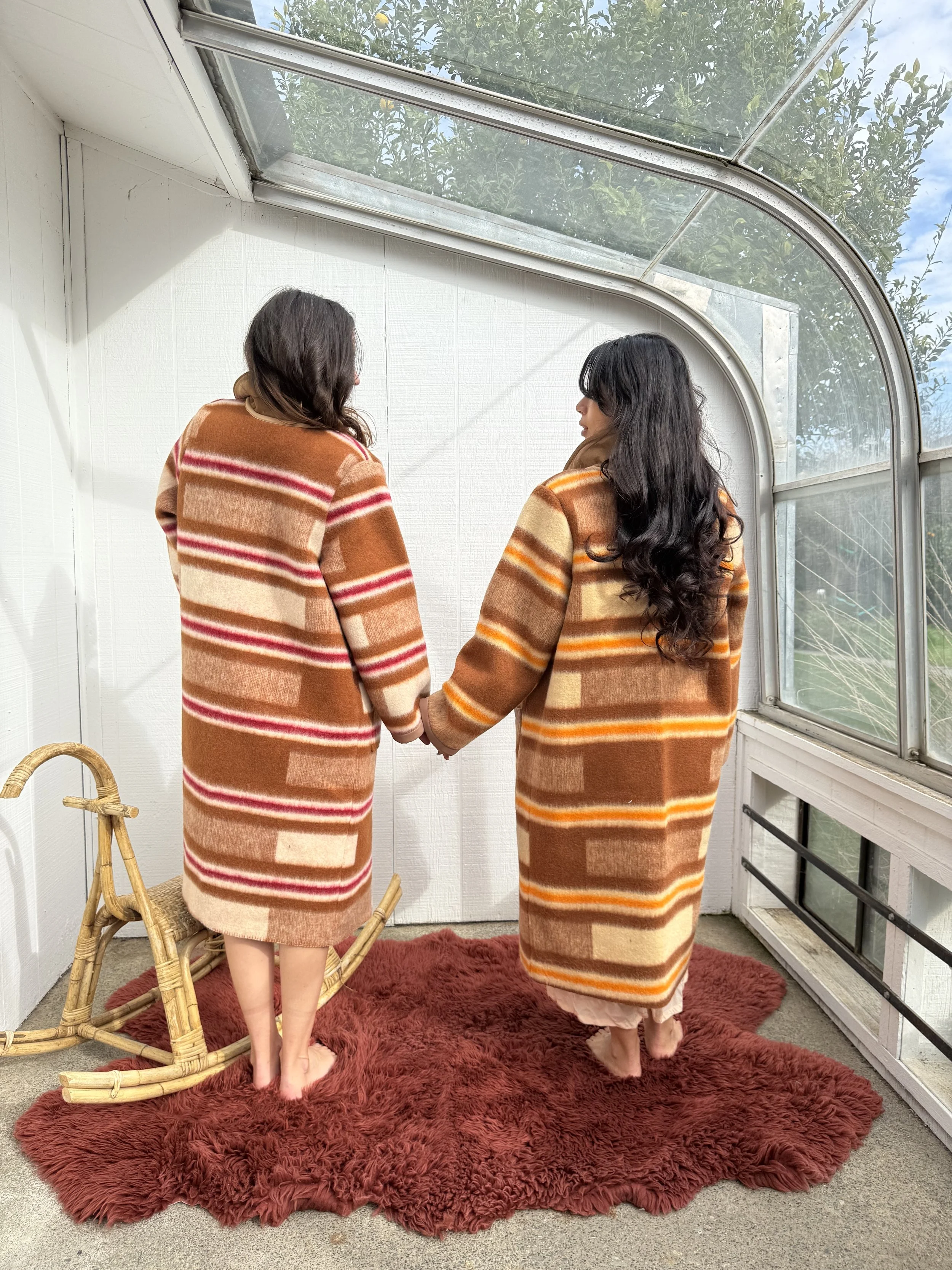 Two women holding hands in a sunlit indoor space with large window, both wearing matching orange, beige, and brown striped coats, standing on a red shaggy rug.