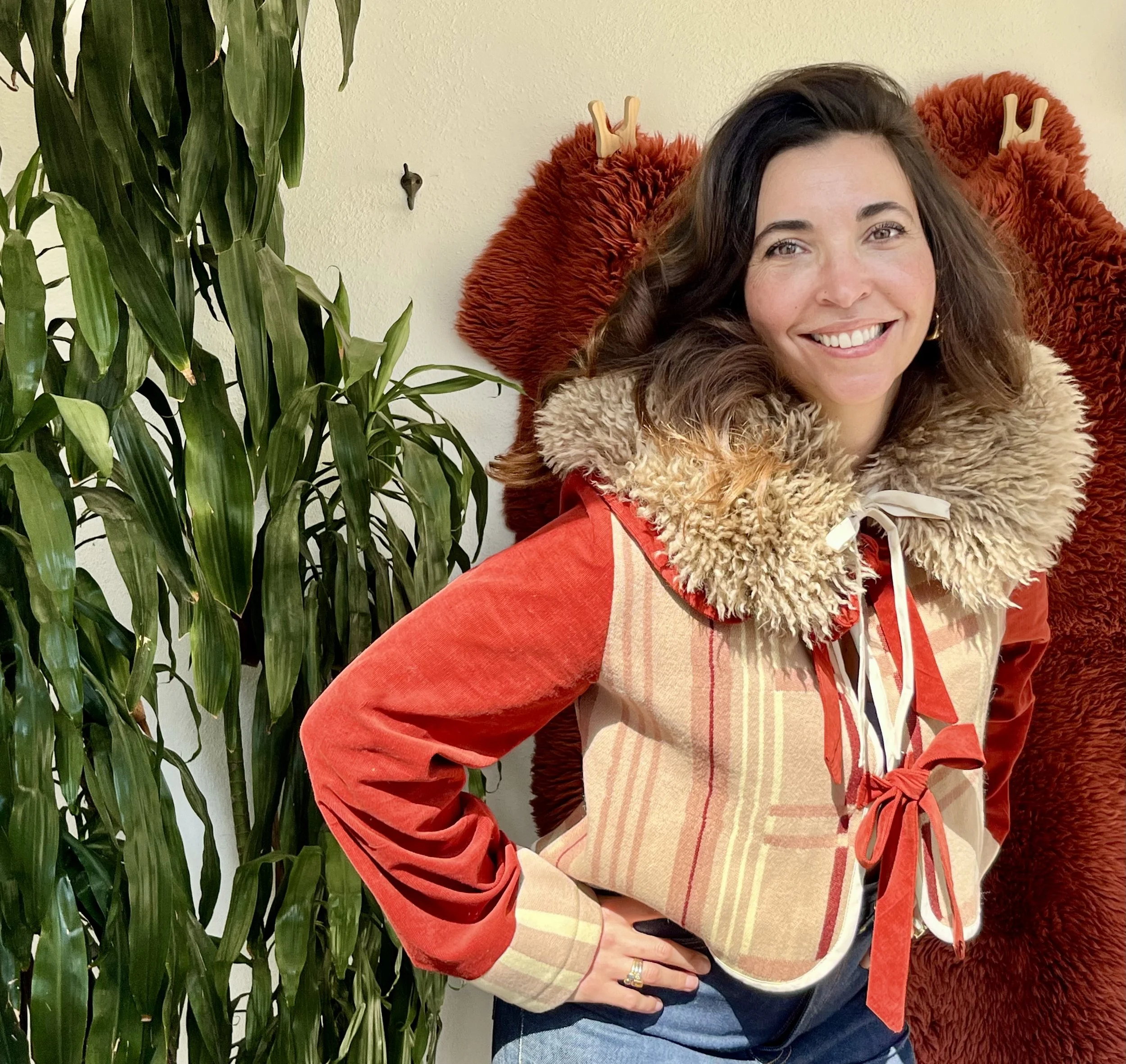A woman with brown hair smiling, wearing a beige and red jacket with a furry collar, standing indoors next to a green plant and red furry objects hanging on the wall.