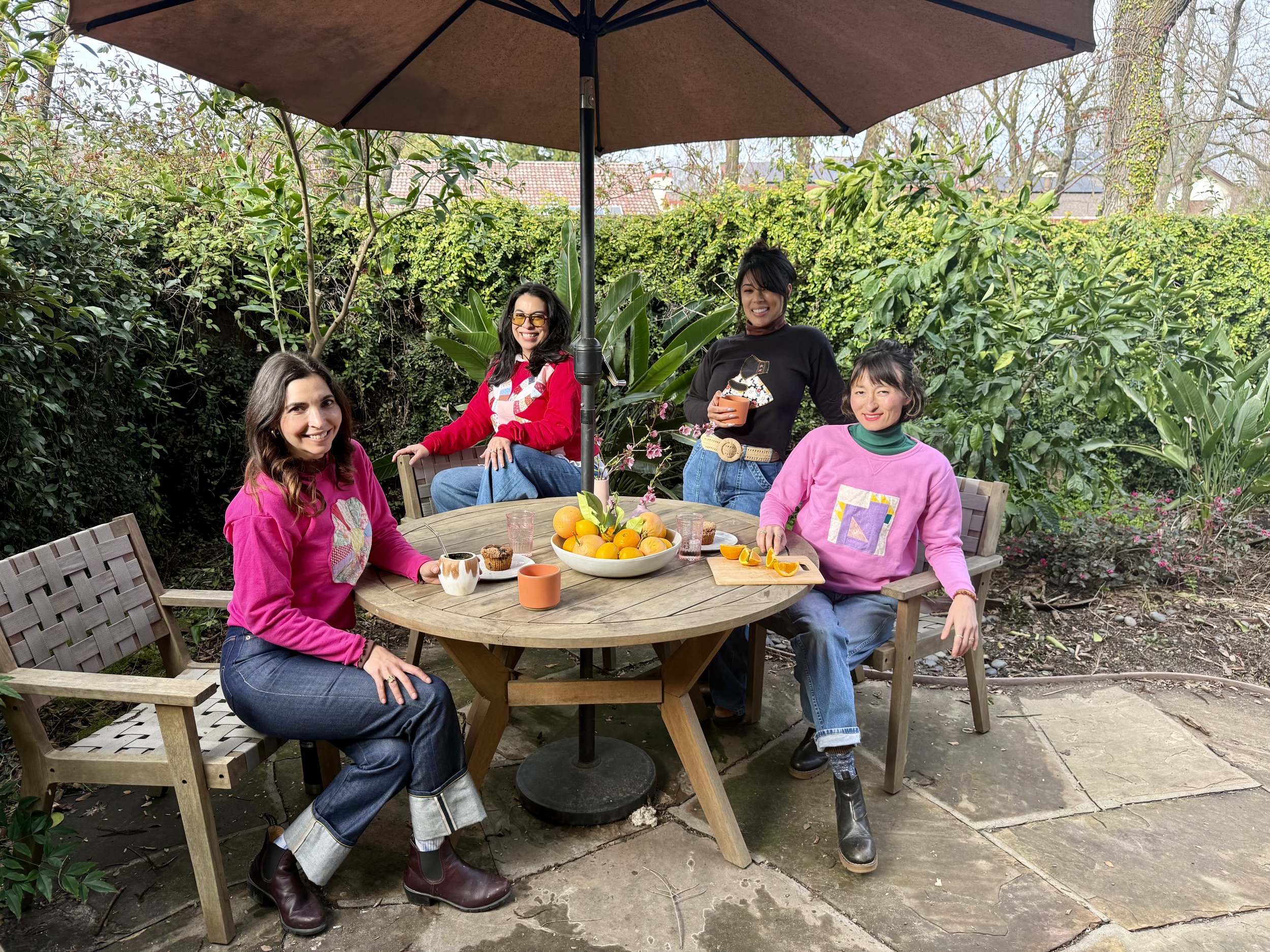 Four women sitting and standing around a wooden outdoor table with fruit, cupcakes, and drinks under a large umbrella in a backyard garden.