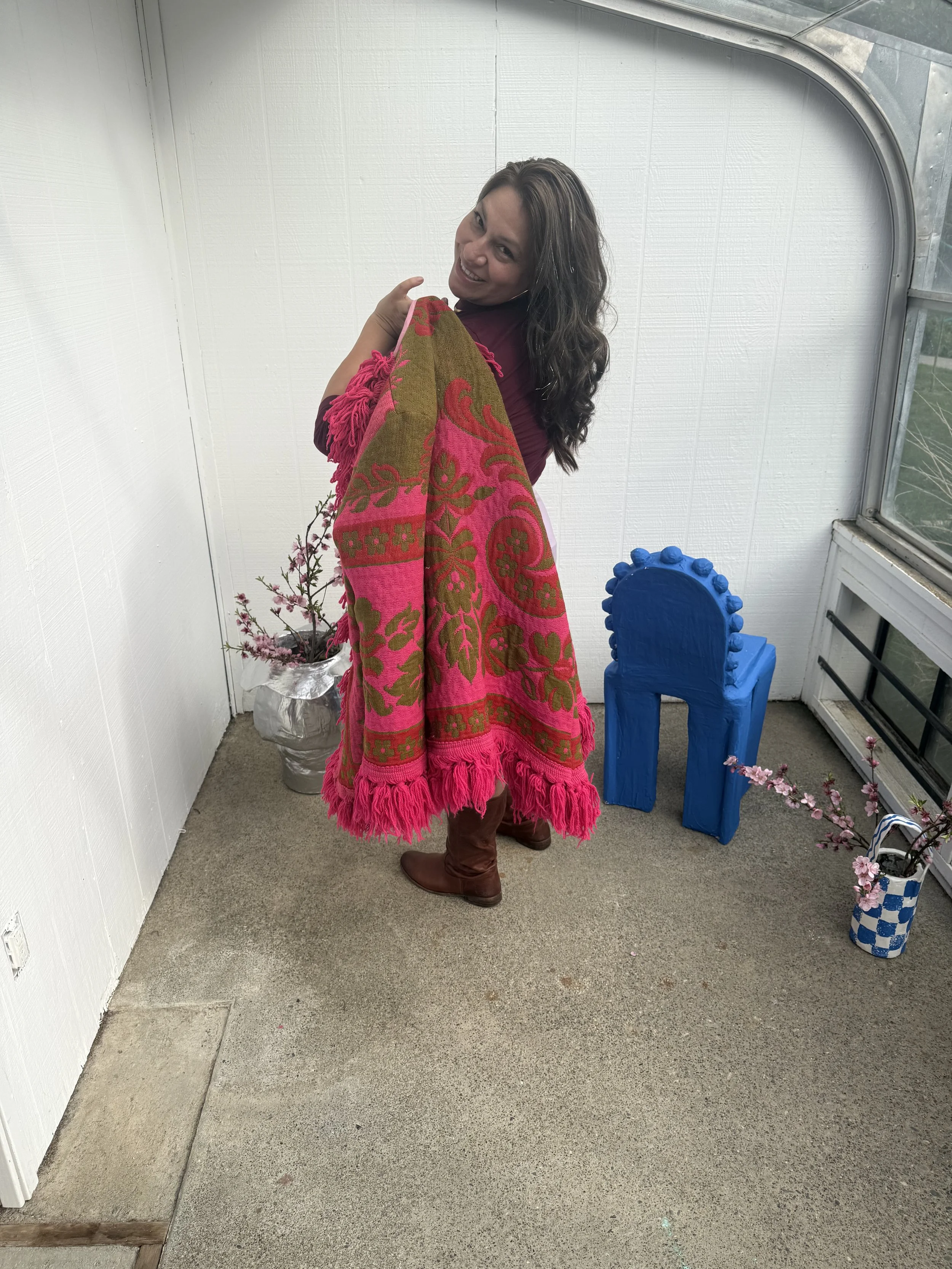 Woman with long dark hair holding a pink and brown patterned blanket, standing on a small enclosed porch with potted fake cherry blossom plants, a bright blue decorative object, and a window with a view outside.