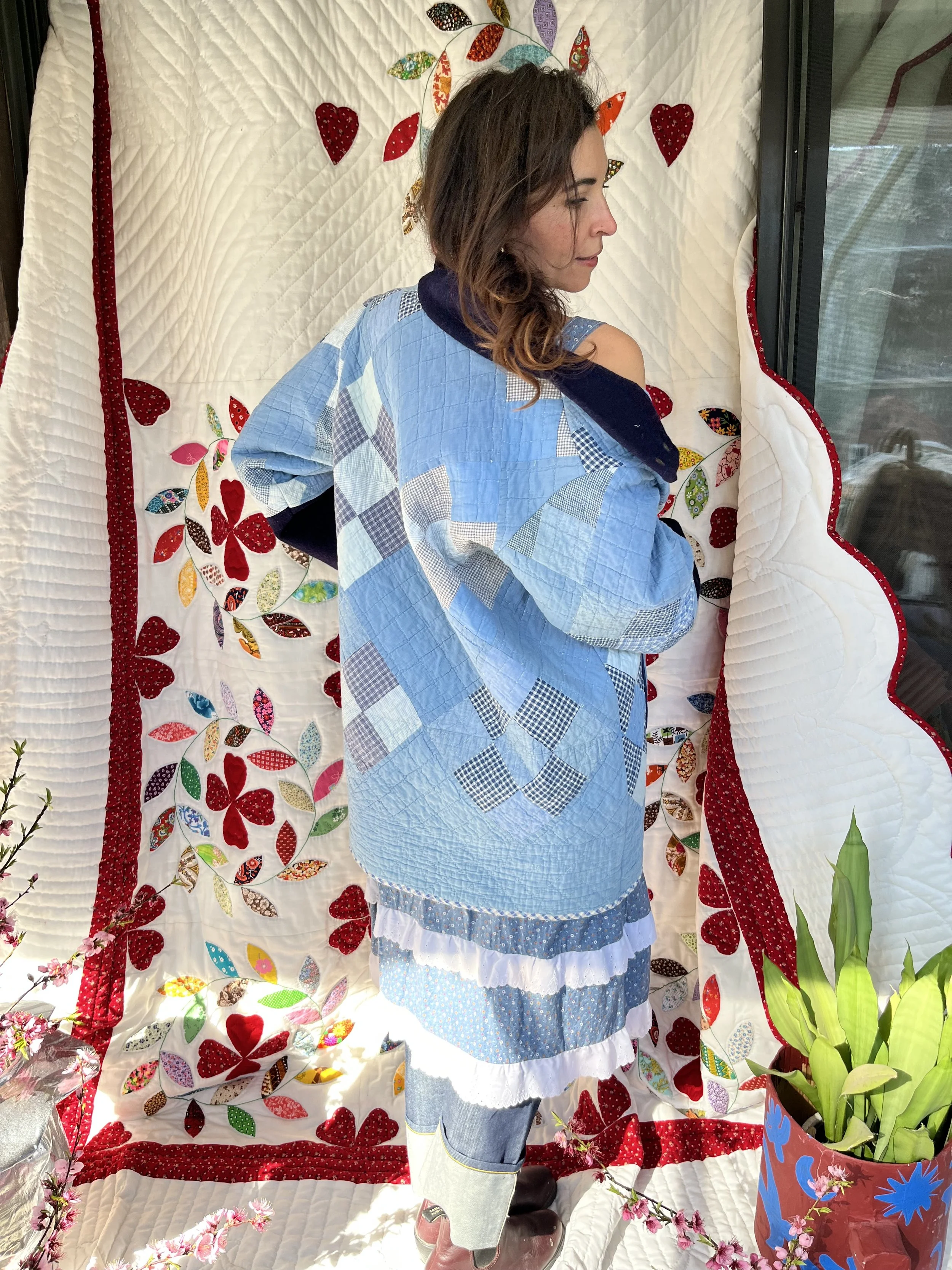 Woman wearing a patchwork blue quilted jacket standing in front of a quilted wall hanging with floral and heart appliqué designs, next to a potted plant with green leaves.