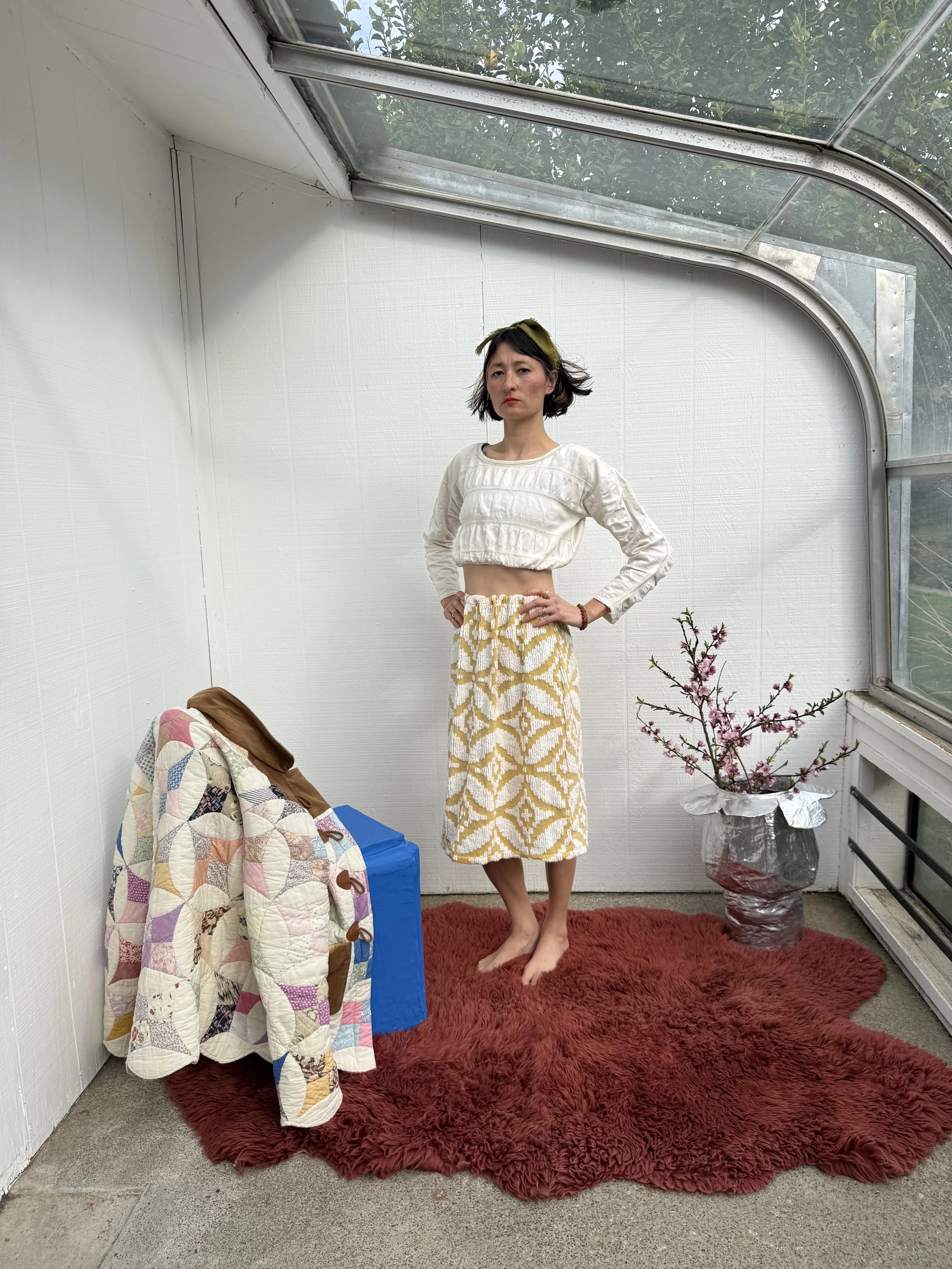 A woman stands barefoot on a red faux fur rug inside a glass-enclosed porch with white walls, posing with hands on hips. She wears a cream-colored, cropped, long-sleeve top and a yellow and white patterned midi skirt. A quilted jacket with pastel pat