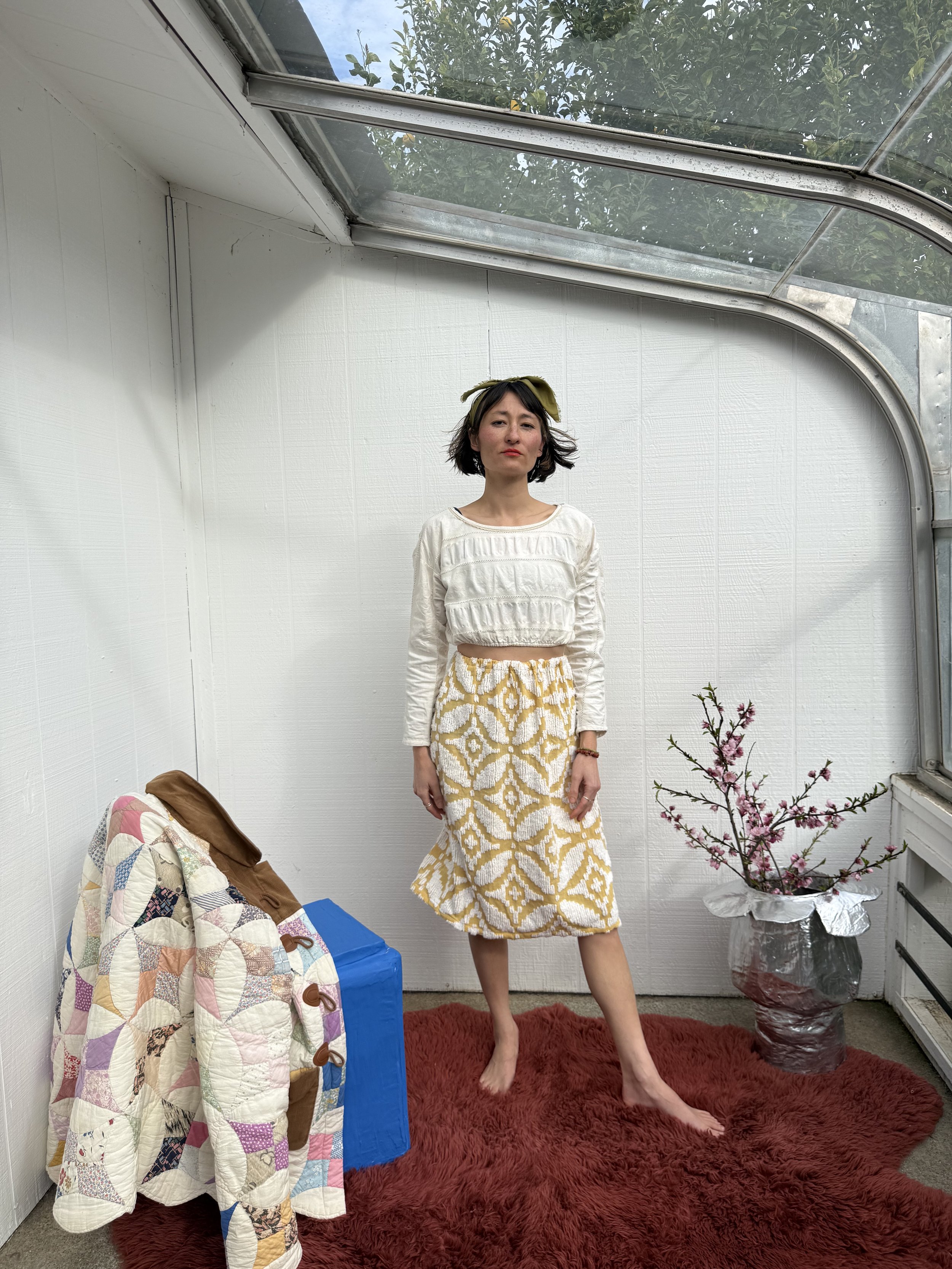 A woman standing barefoot on a red rug inside a small greenhouse or sunroom with white walls, a large glass roof, a flowering branch in a silver vase, a multicolored quilt draped over a blue stool, and some greenery visible through the glass ceiling.