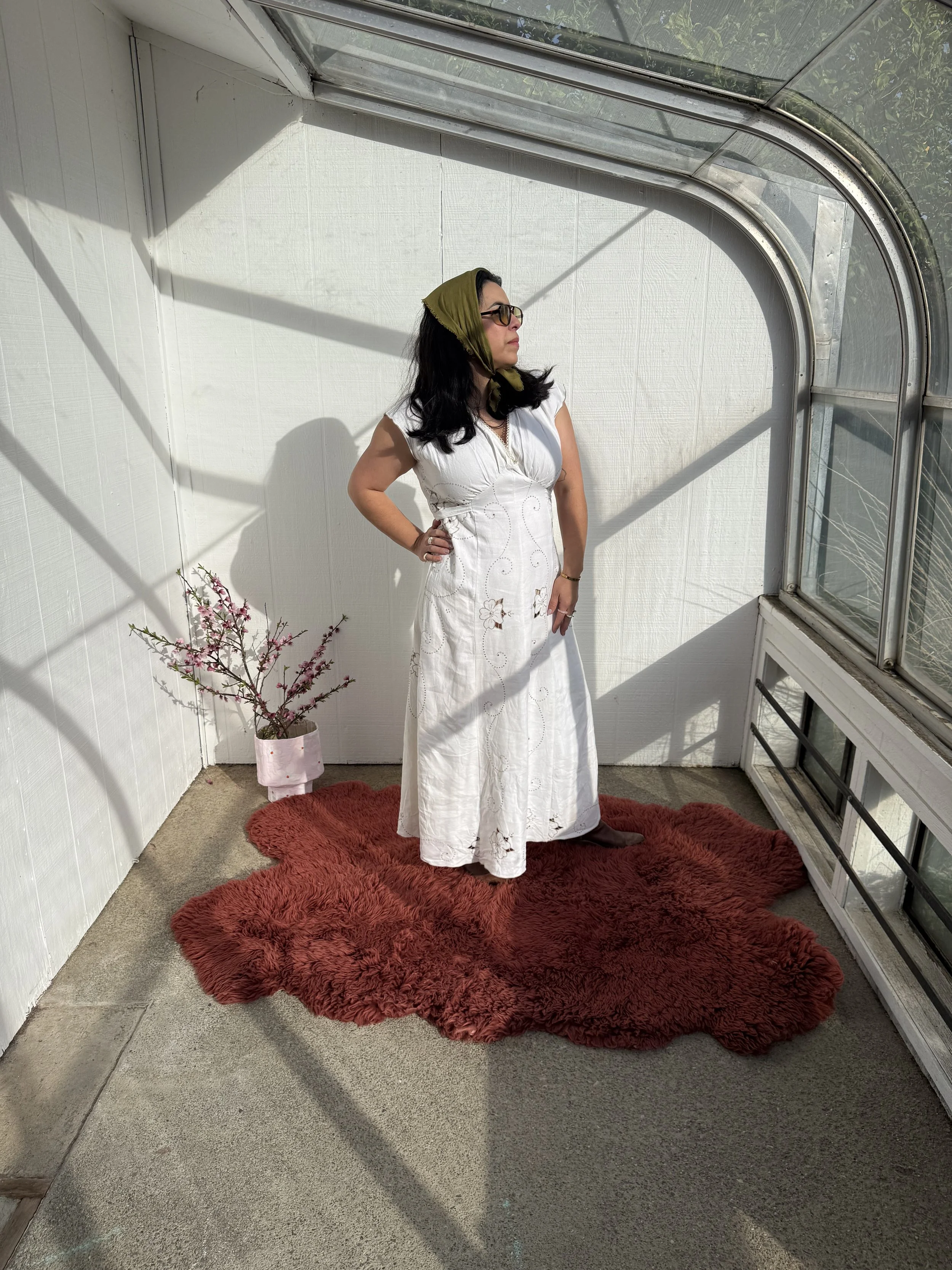 A woman with black hair, wearing glasses, a white dress, and a green headscarf, standing inside a sunlit greenhouse on a red fluffy rug, with a potted flowering plant nearby.