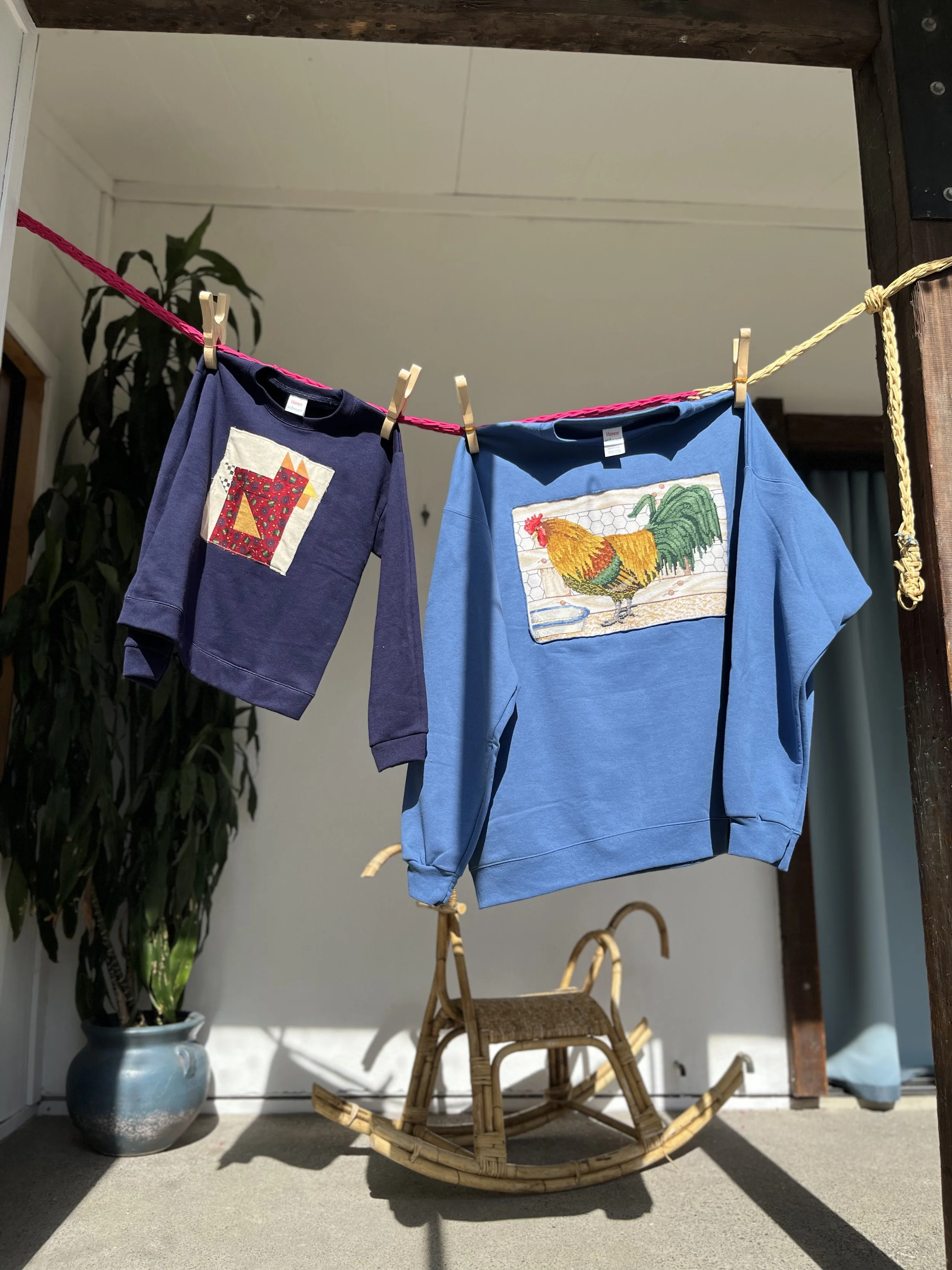 Children's clothing hanging on a clothesline with clothespins, two sweatshirts featuring animal and nature-themed embroidery, outdoors with a plant and a wicker rocking chair in the background.