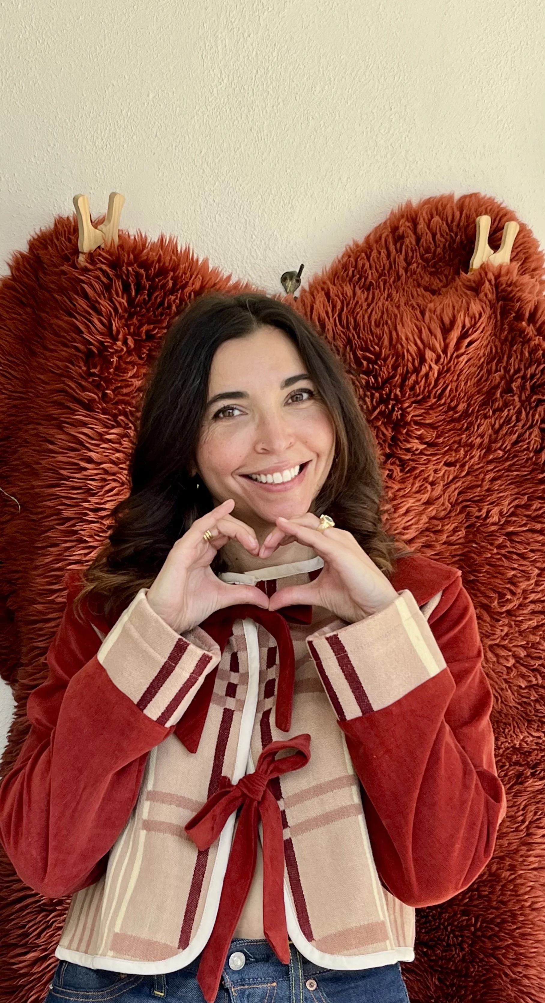 A woman with brown hair smiling, making a heart shape with her hands, sitting in front of a large red furry butterfly-shaped chair with two maple leaf-shaped wooden hooks, and a small bird perched on her head.