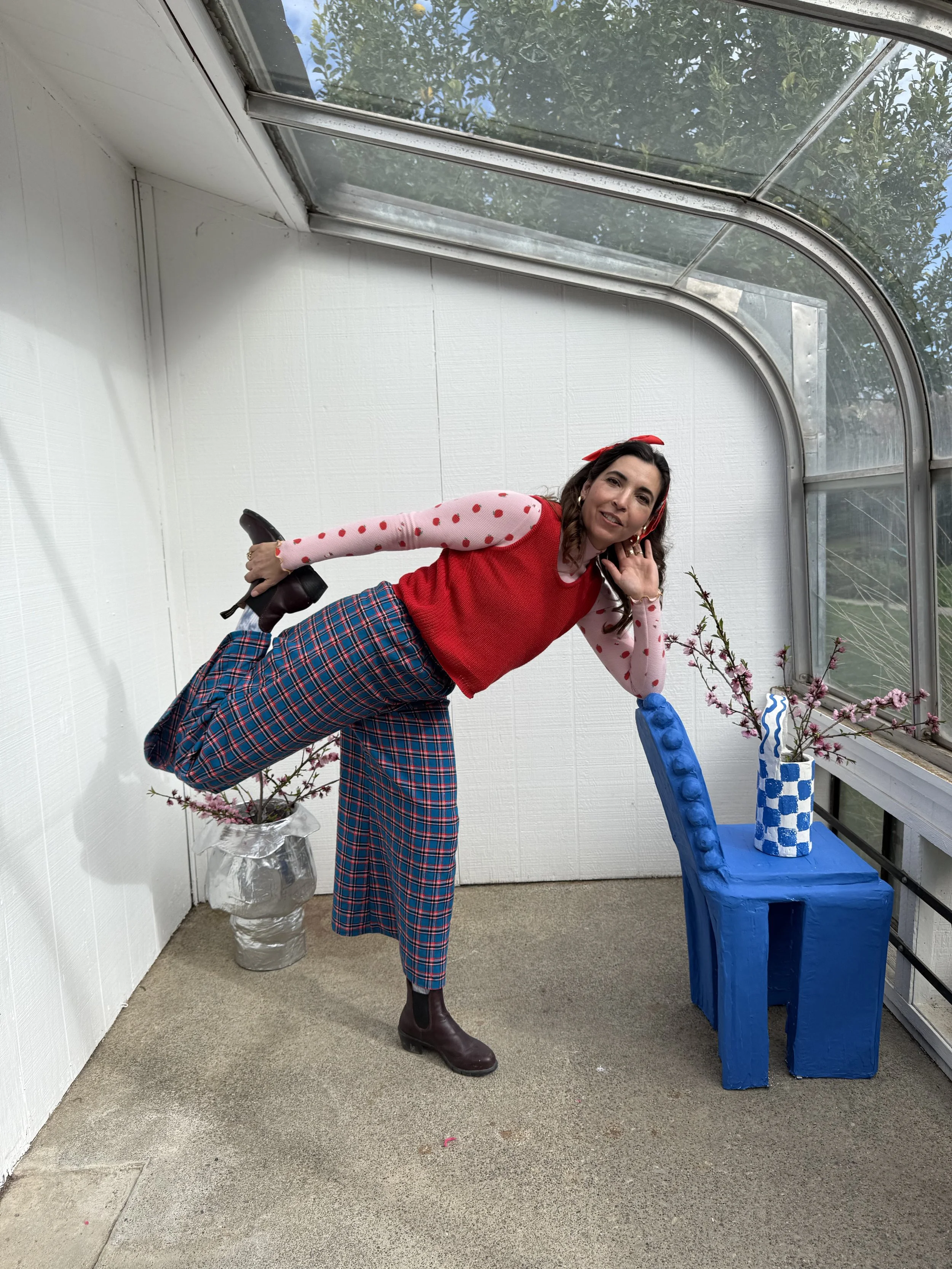 Woman in plaid pants, red sweater vest, and boots standing on one leg, with her other leg held behind her, inside a bright greenhouse with a blue chair, pink blossom branches, and a white wall.