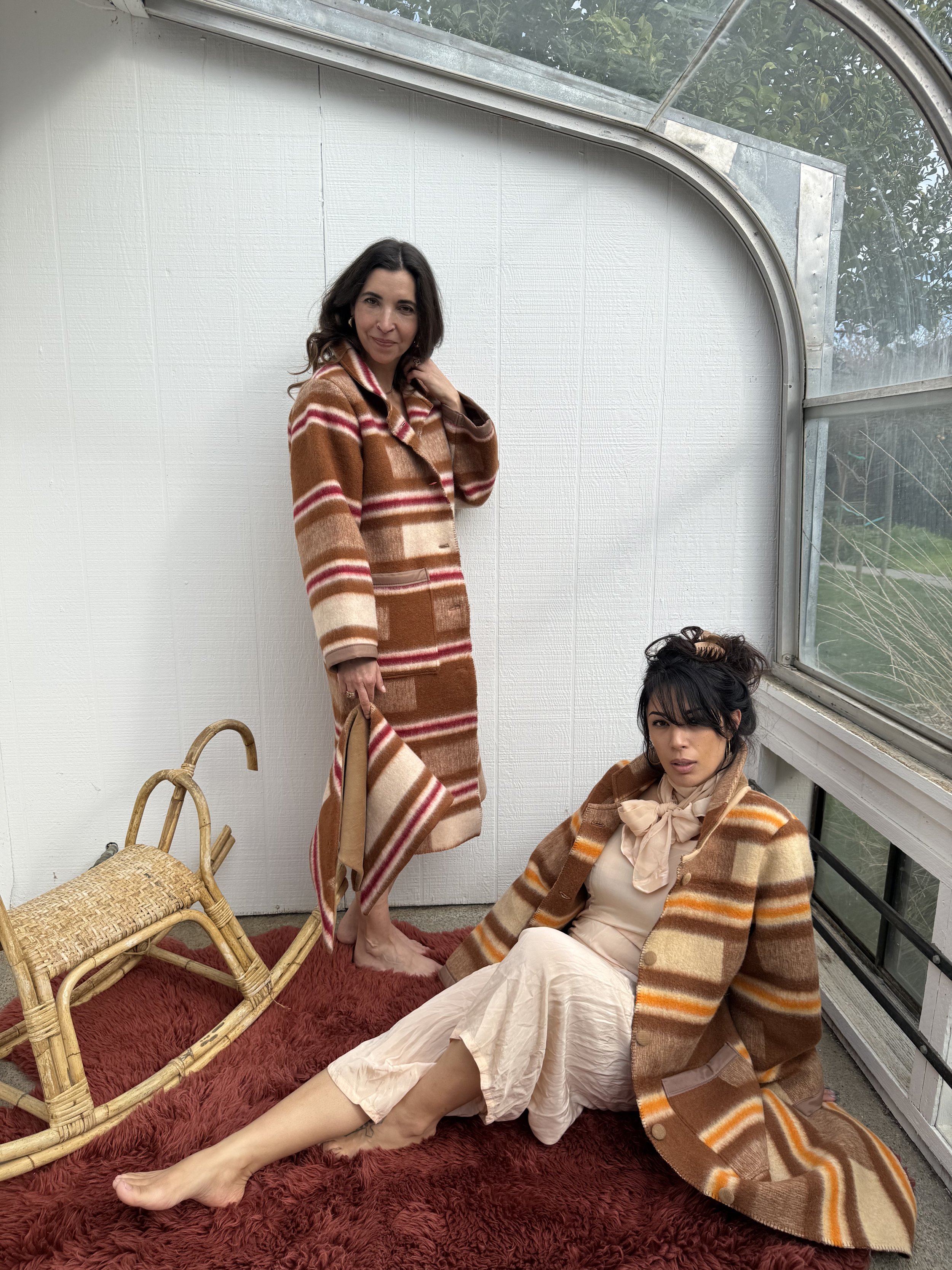Two women wearing brown and beige striped coats in a sunlit room with white walls and large windows. One woman is sitting on a red shaggy rug, and the other is standing beside her, holding a matching coat.