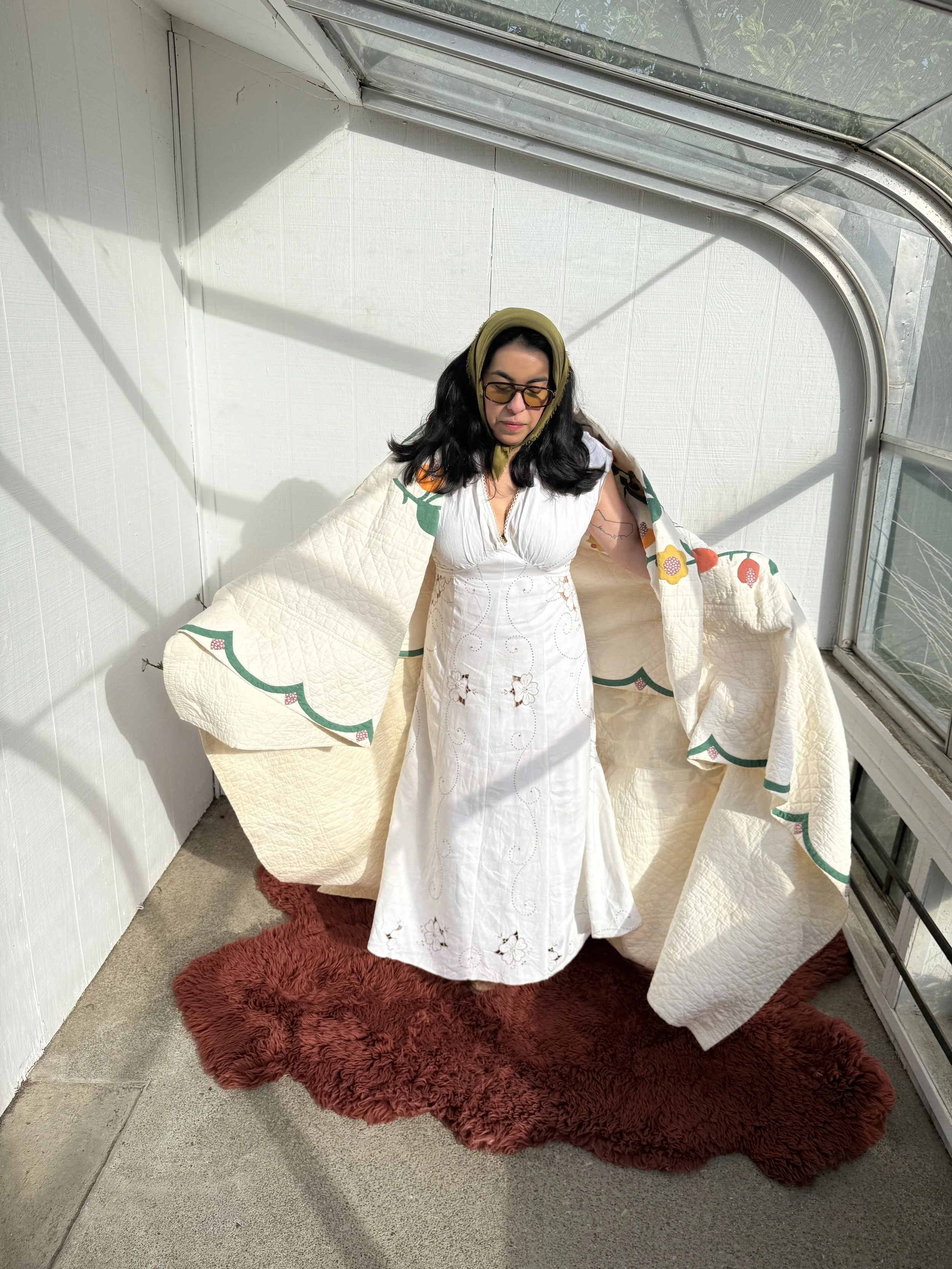Woman wearing a white dress with embroidered floral details and a quilted cream and green patterned shawl, standing on a red shaggy rug inside a sunlit room with glass walls.