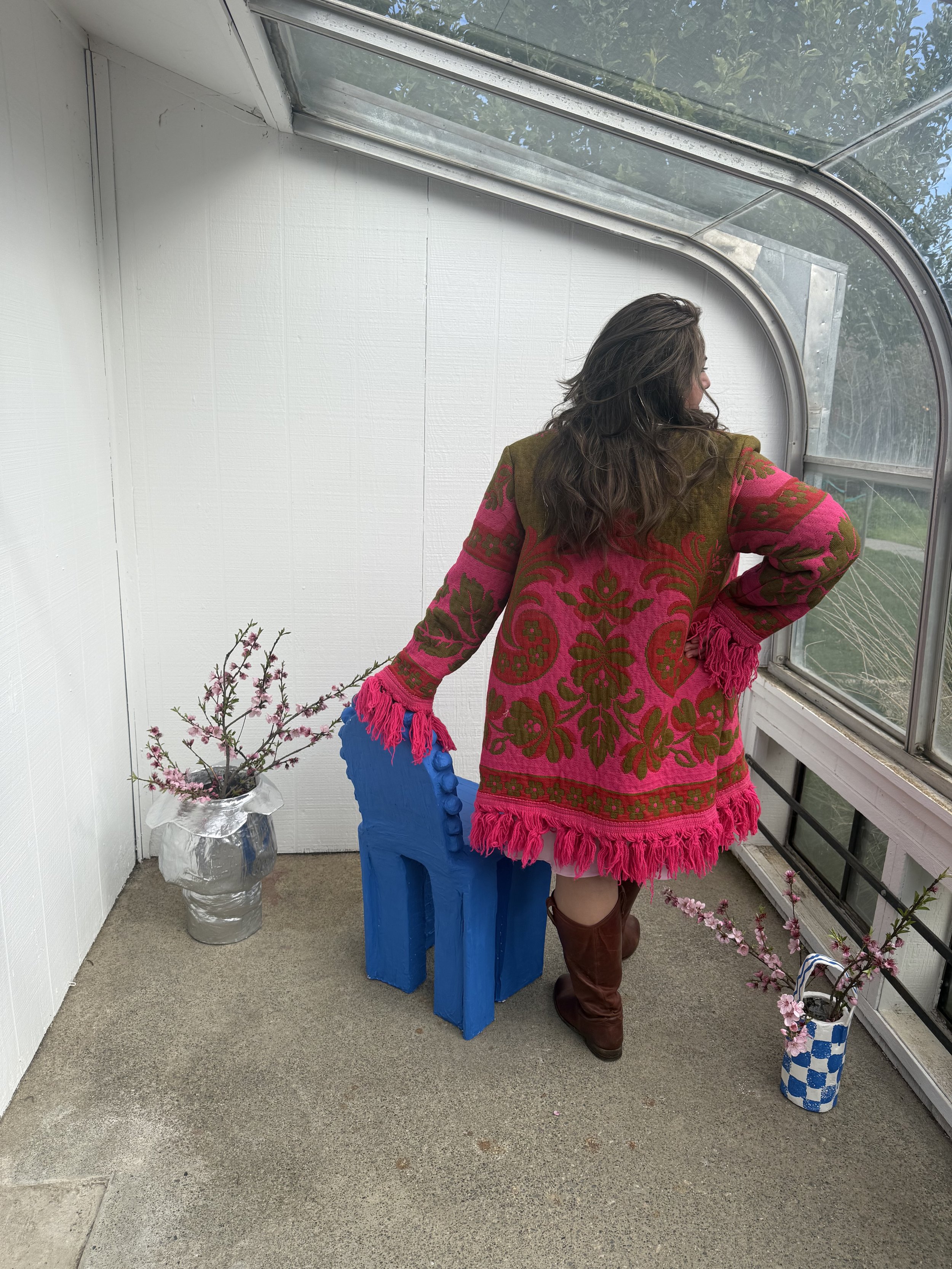 A woman with long brown hair, wearing a pink and brown patterned coat with fringe, is standing in front of a window on a small enclosed porch or sunroom. She faces away and looks out the window. There are two planters with pink flowering branches, on