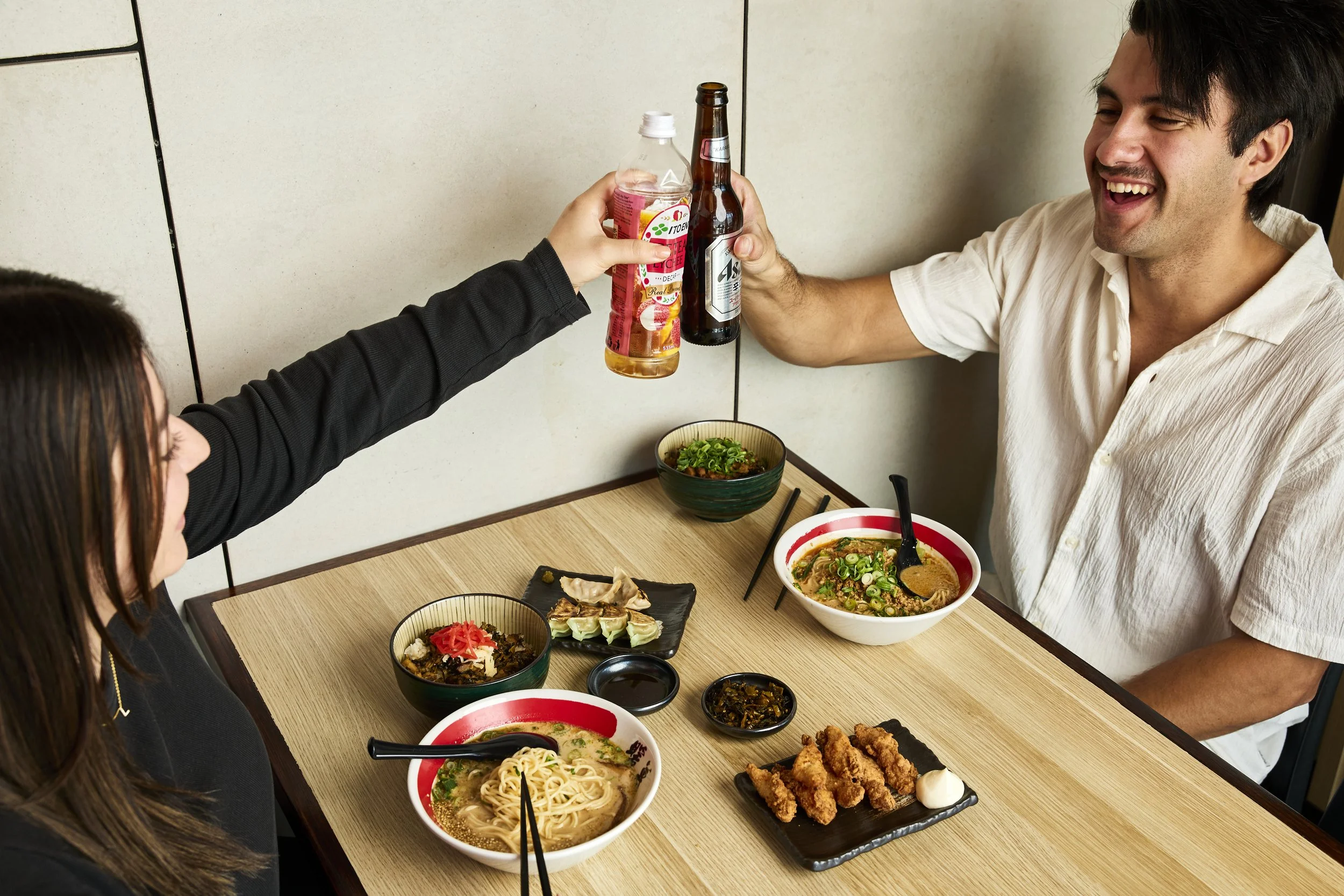 Zwei Menschen beim Essen, die auf ein Glas anstoßen, im Vordergrund verschiedener japanischer Gerichte auf dem Tisch, darunter Ramen und Gyoza.
