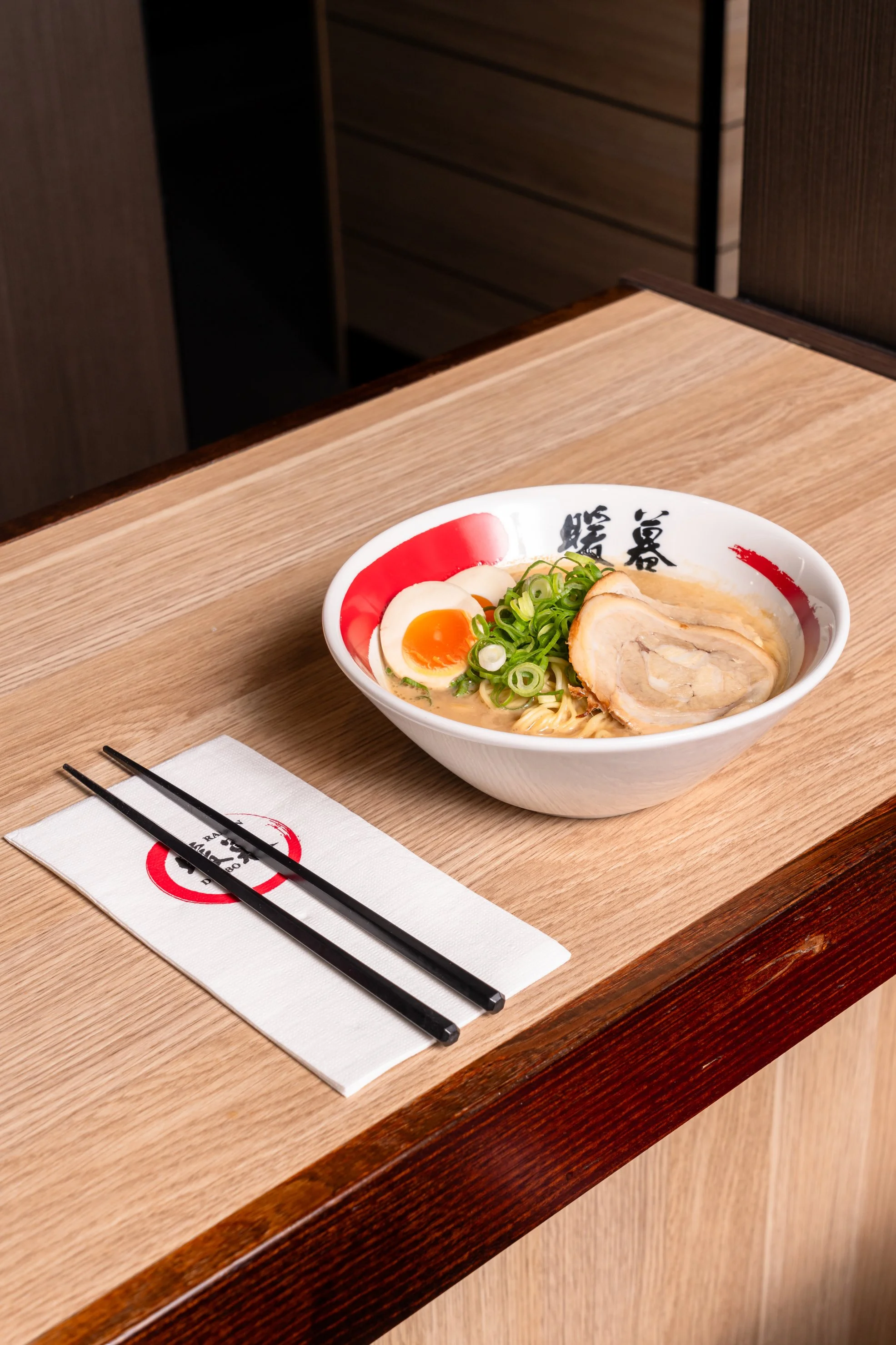 Schüssel mit Ramen-Nudeln, halbiertes Ei, Scheiben von Schweinefleisch und Frühlingszwiebeln auf einem Holztisch mit Essstäbchen und Papierserviette.