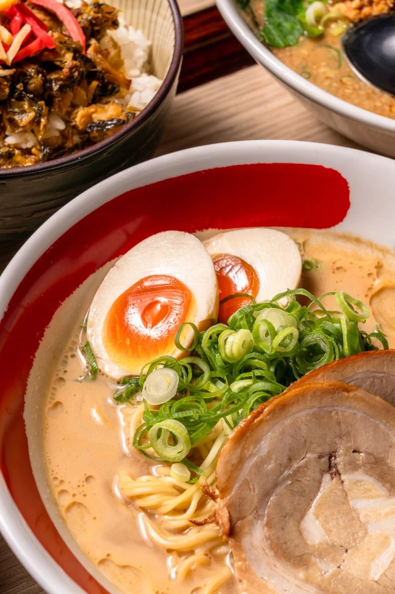 Ein Schüssel mit Ramen-Nudeln, weich gekochtem Ei, Frühlingszwiebeln und Schweinefleisch, daneben zwei weitere Schüsseln mit Reis und Suppe.