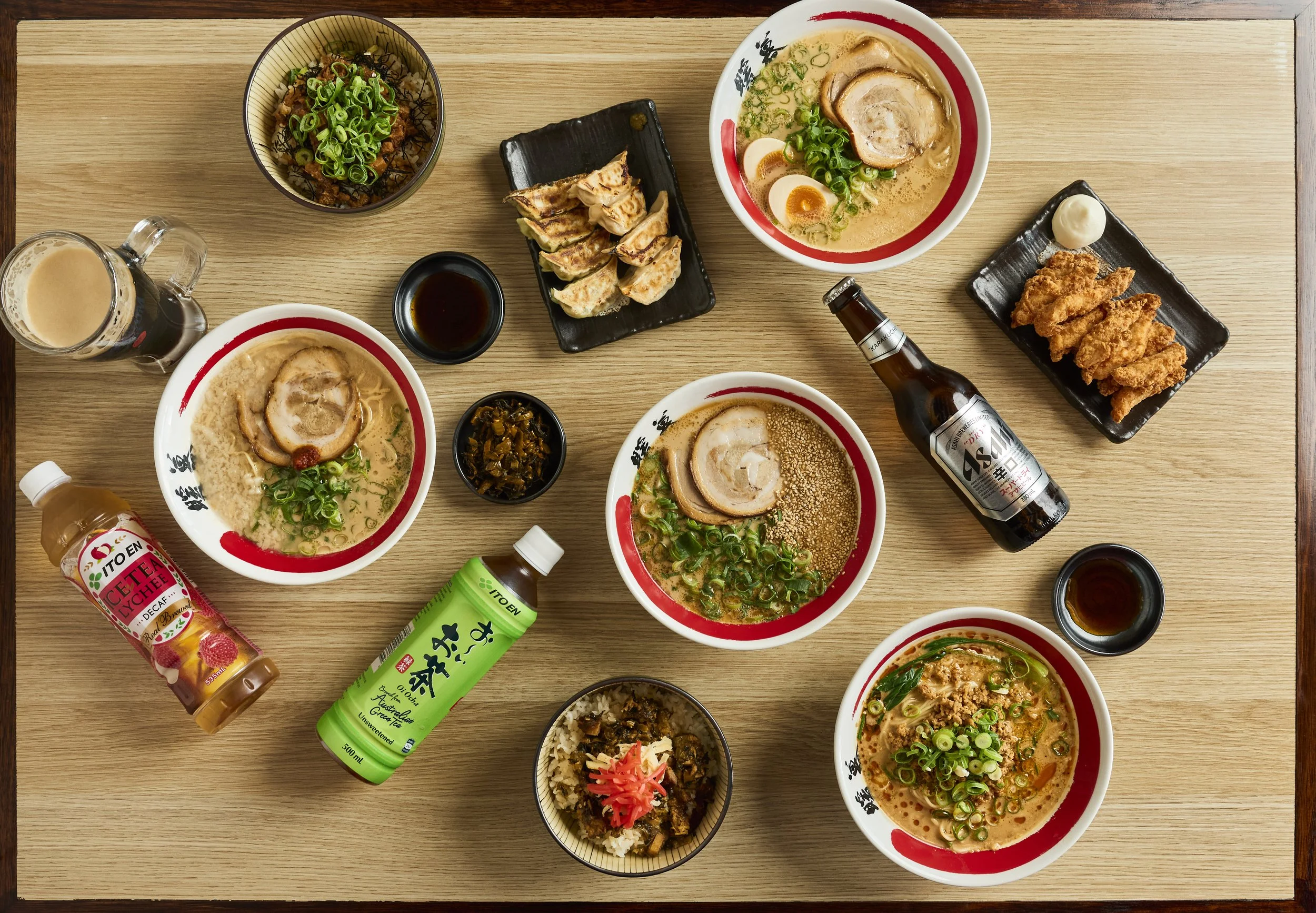 Verschiedene Schüsseln mit Ramen-Nudeln, Gyoza, frittiertes Hühnchen, Soßen und Getränke auf einem Holztisch