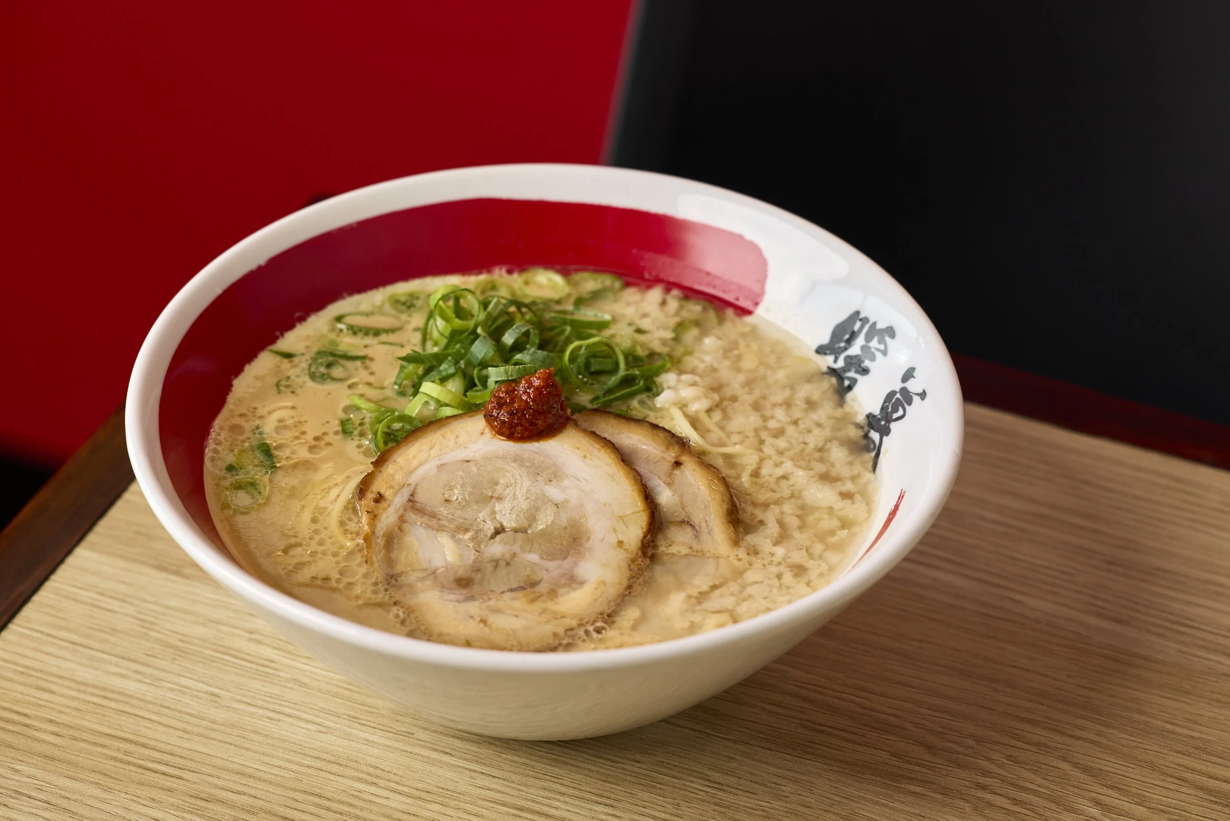 Schüssel mit Ramen-Nudeln, Schweinefleisch, grünen Zwiebeln und roter Paste auf einer Holzplatte.