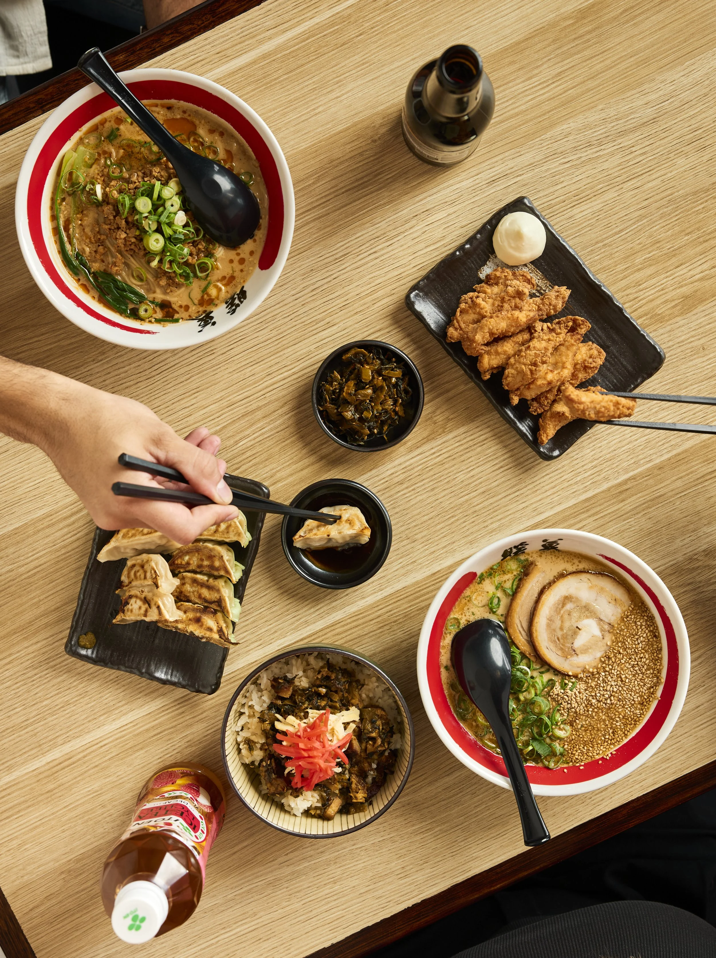 Verschiedene japanische Gerichte auf einem Holztisch, darunter Ramen-Nudeln, Gyoza, fried chicken, Reis mit Toppings und Soßen, sowie eine Flasche Sojasoße.