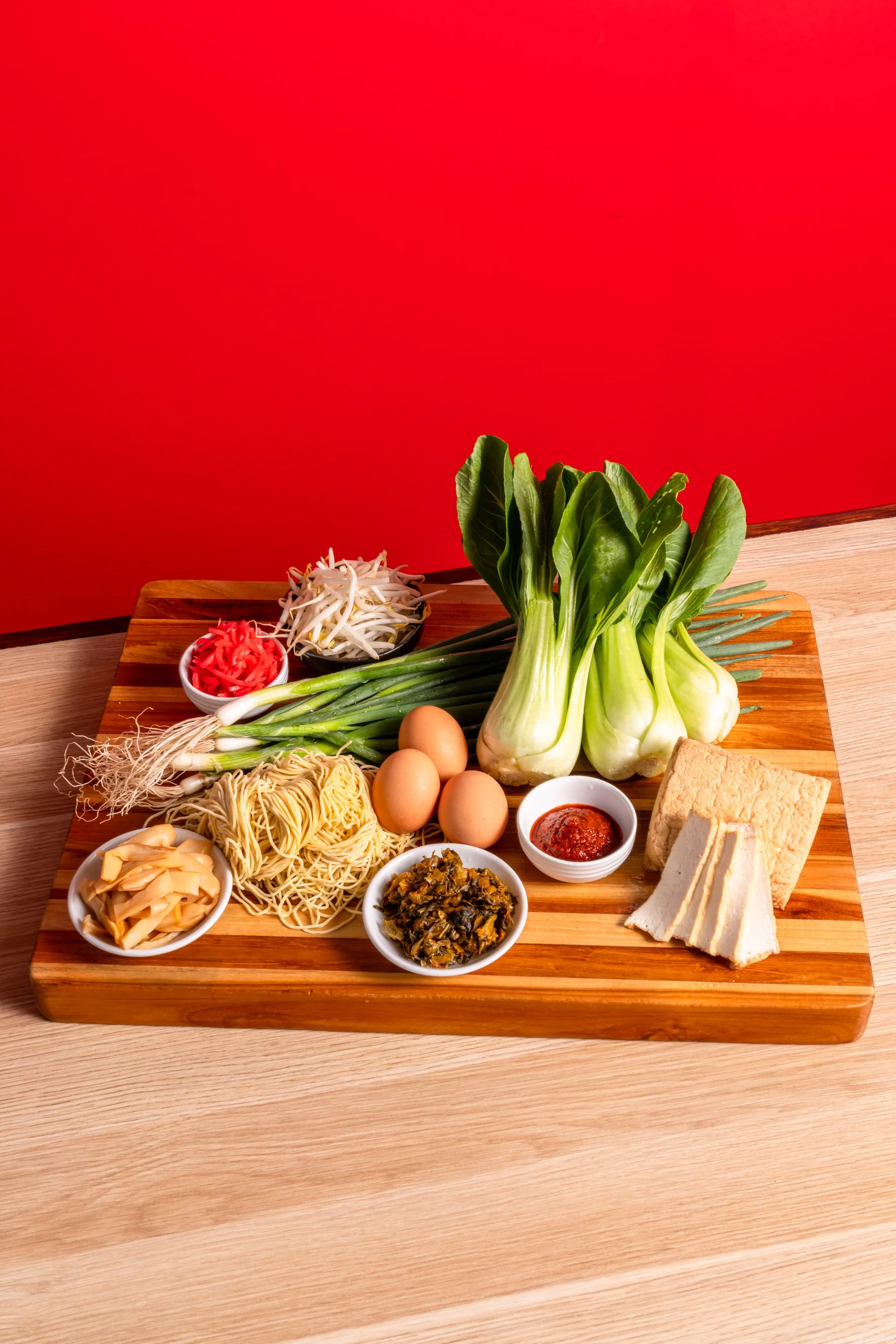 Verschiedene Zutaten für ein Gericht, darunter Eier, Gemüse, Nudeln, Soßen und Tofu, auf einem Holzbrett.