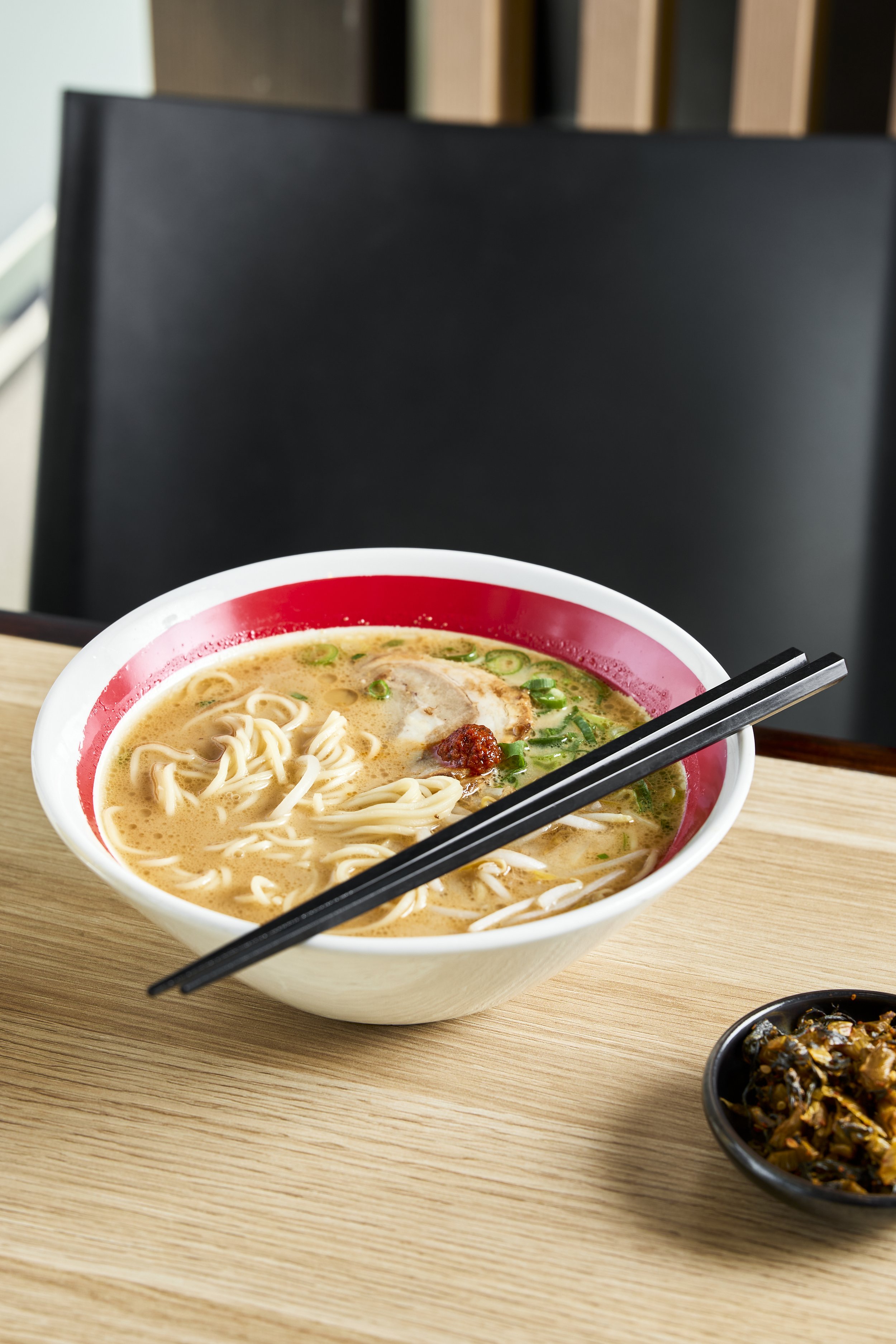 Schüssel mit Ramen-Nudeln, grünen Zwiebeln, Huhn und Chilli Paste, dazu ein kleines schwarzes Schälchen mit Gemüse, auf einem Holztablett.